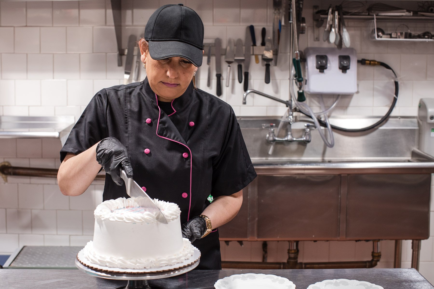 A female pastry chef in a black uniform and cap decorates a white cake with icing in a commercial kitchen.