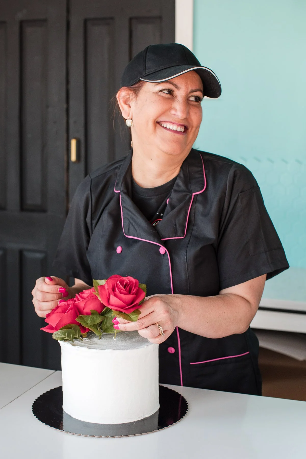 A woman in a black chef's coat with pink piping, wearing a black cap, smiling and holding a bouquet of pink roses, standing next to a white frosted cake with pink roses on top.