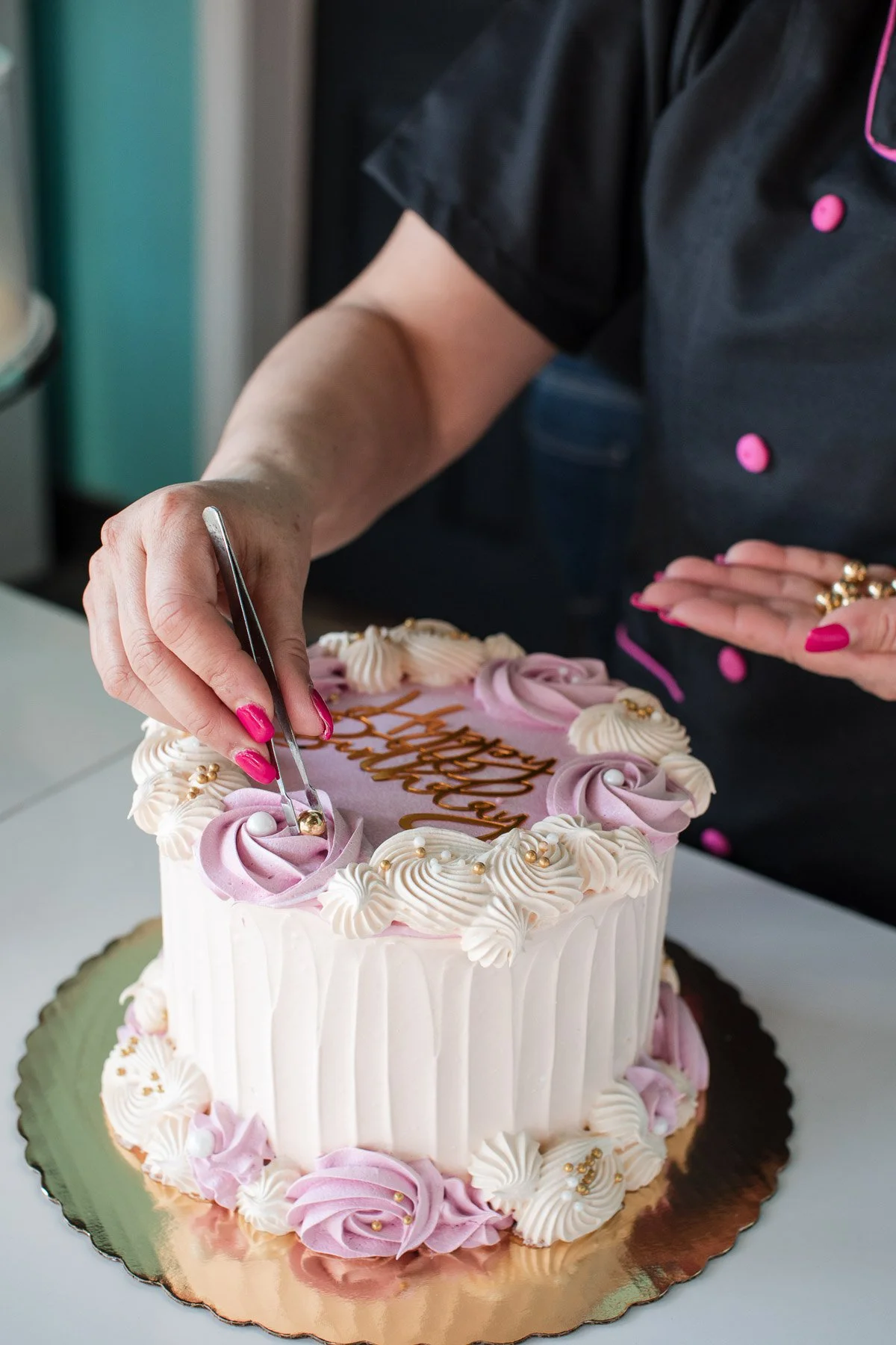 A person in black with pink buttons decorating a pink and white cake with pink and white frosting roses and gold accents, using tweezers to place a gold decoration