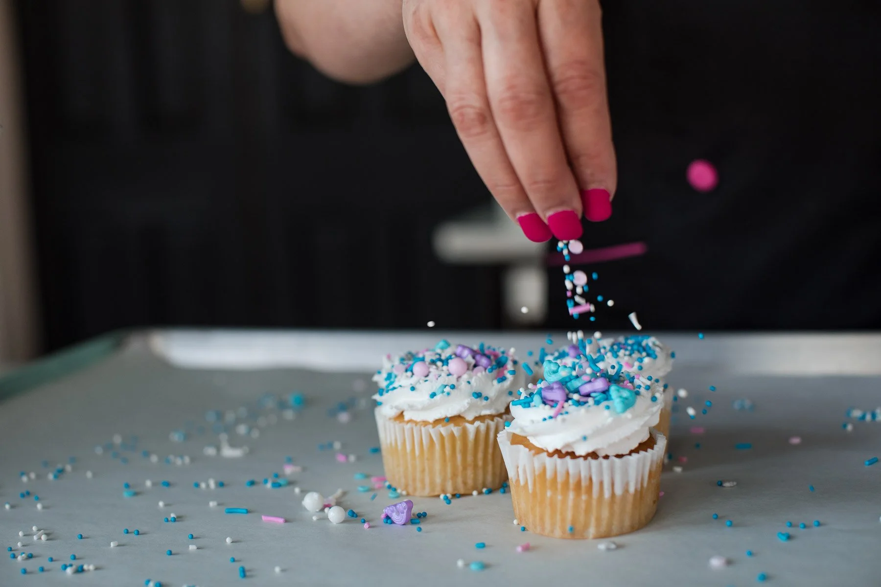 Hand with pink painted nails sprinkling colorful sprinkles onto cupcakes with white frosting and pastel-colored decorations.