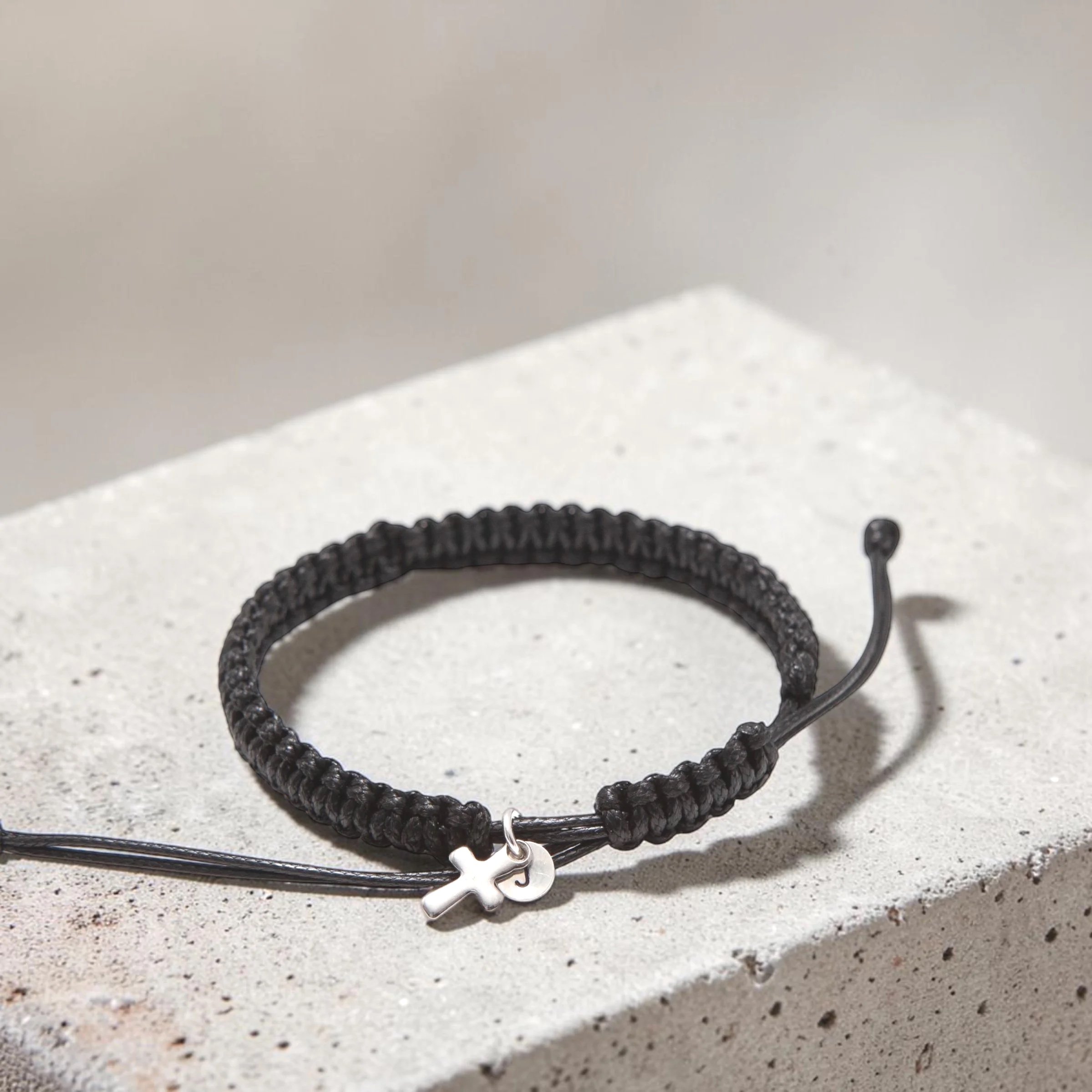 A black braided rope bracelet with a small silver cross charm on a white textured surface.