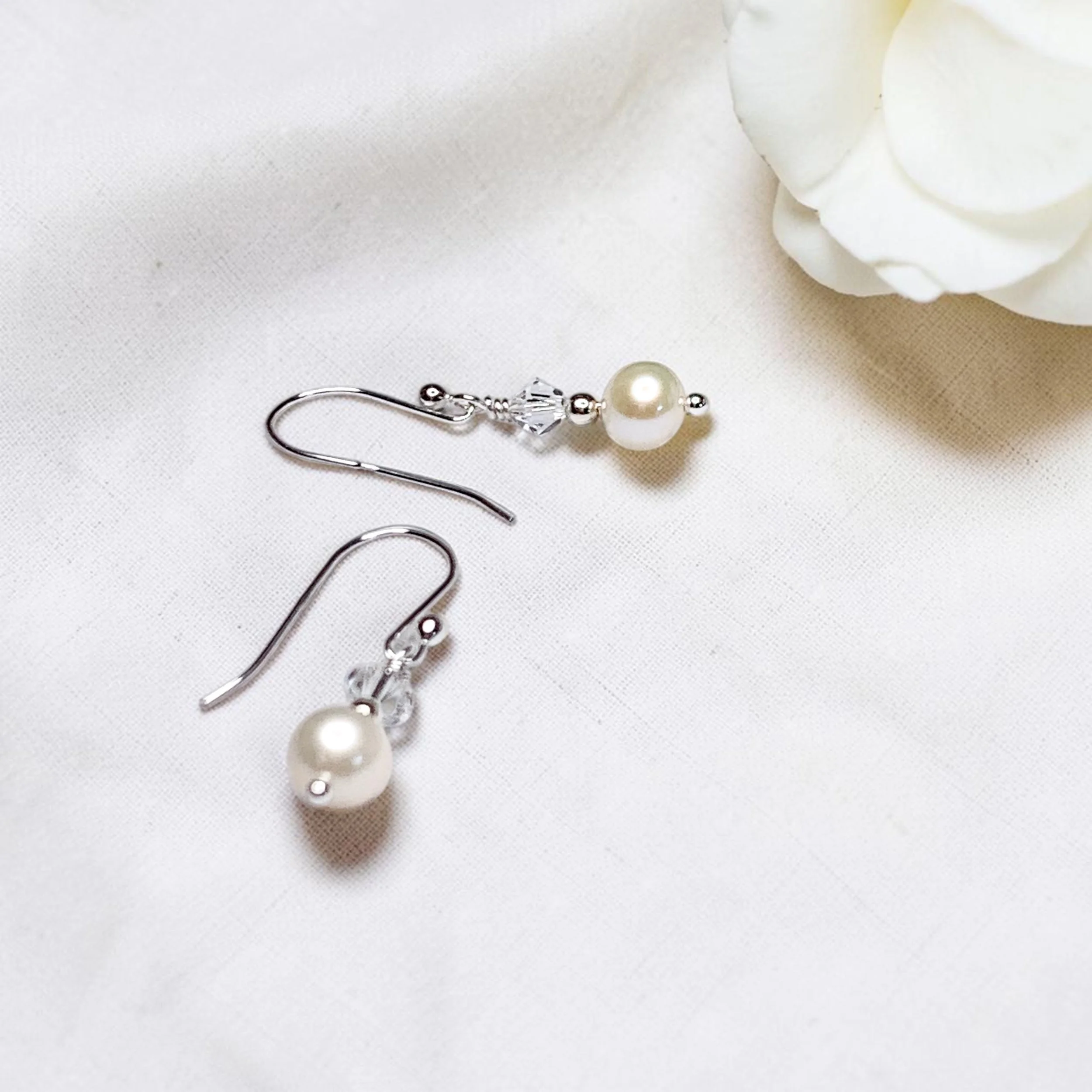 A pair of pearl earrings with crystal accents and metal hooks are placed on a white surface near a white flower.
