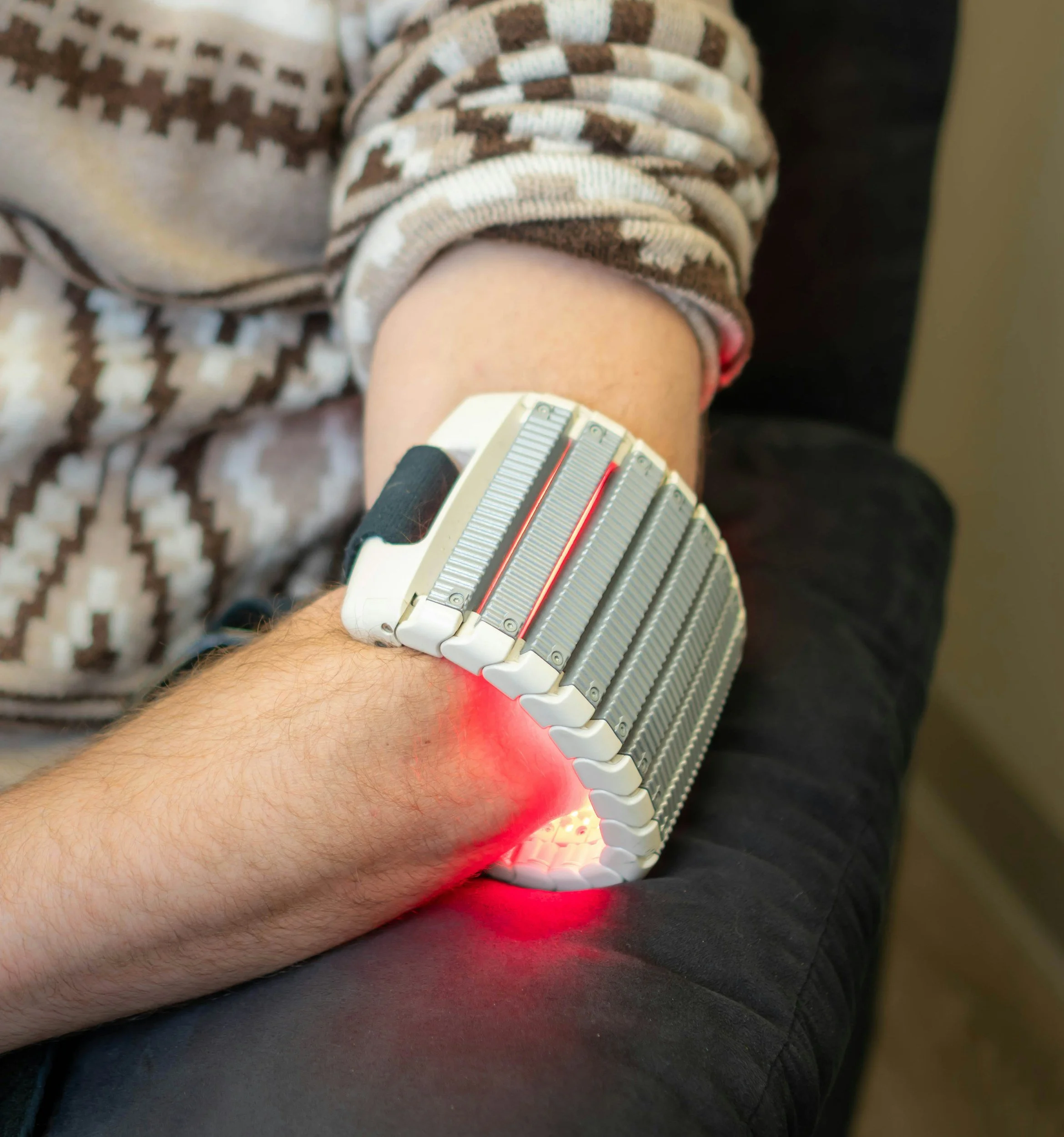 Close-up of a person's arm resting on a black surface, with a futuristic device emitting red light attached around their forearm. The device has strips and wires, and the person is wearing a patterned sweater with beige and brown colors.