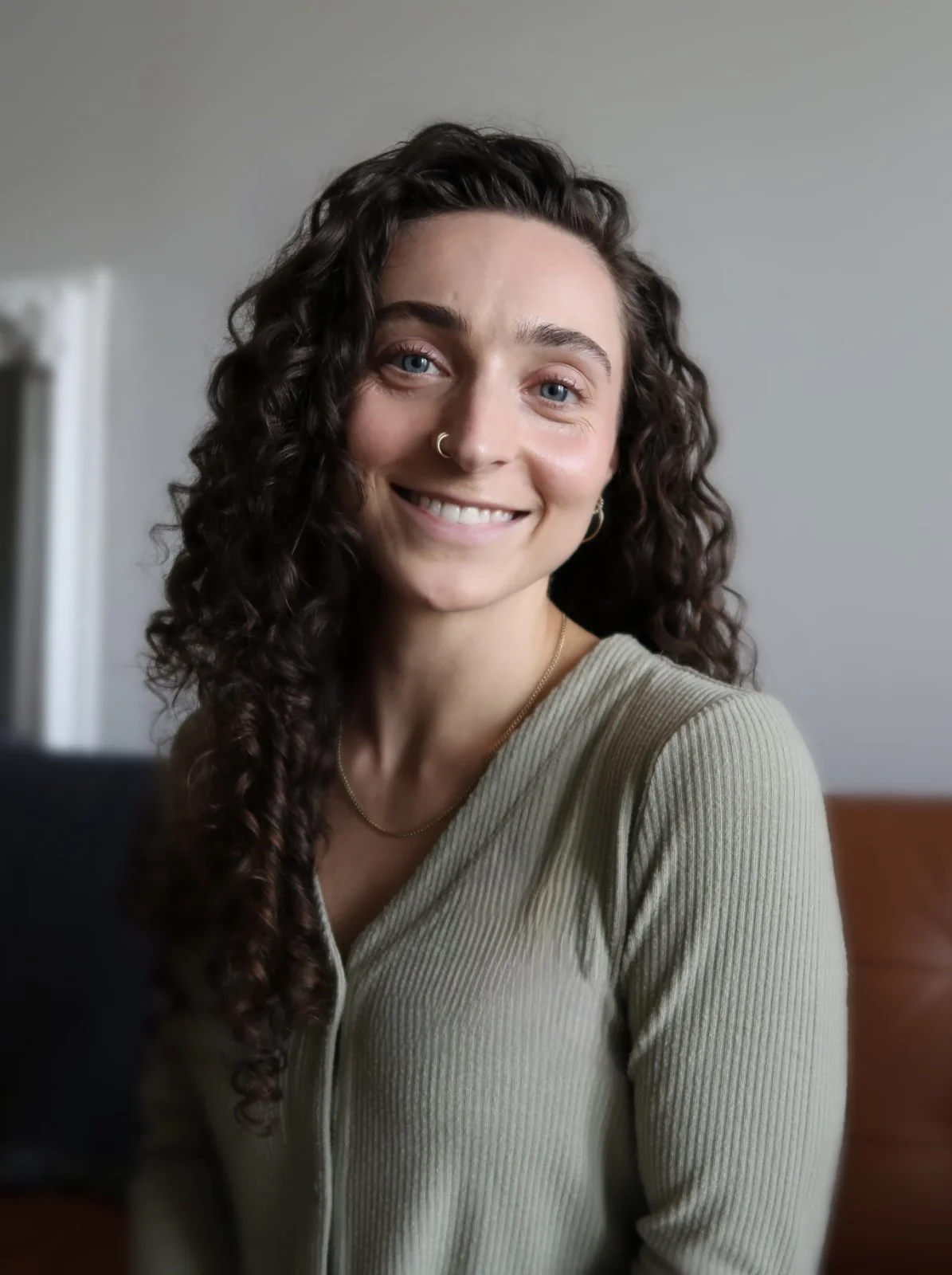 A woman with long, dark, curly hair, blue eyes, and a nose ring, smiling at the camera indoors.