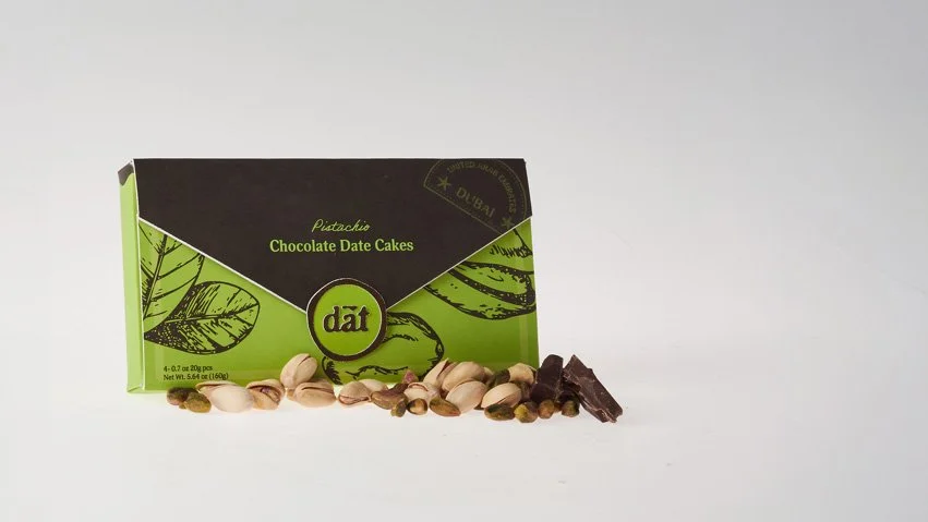 Green and black box of Dāṭ pistachio chocolate date cakes with pistachios and chocolate pieces in front.