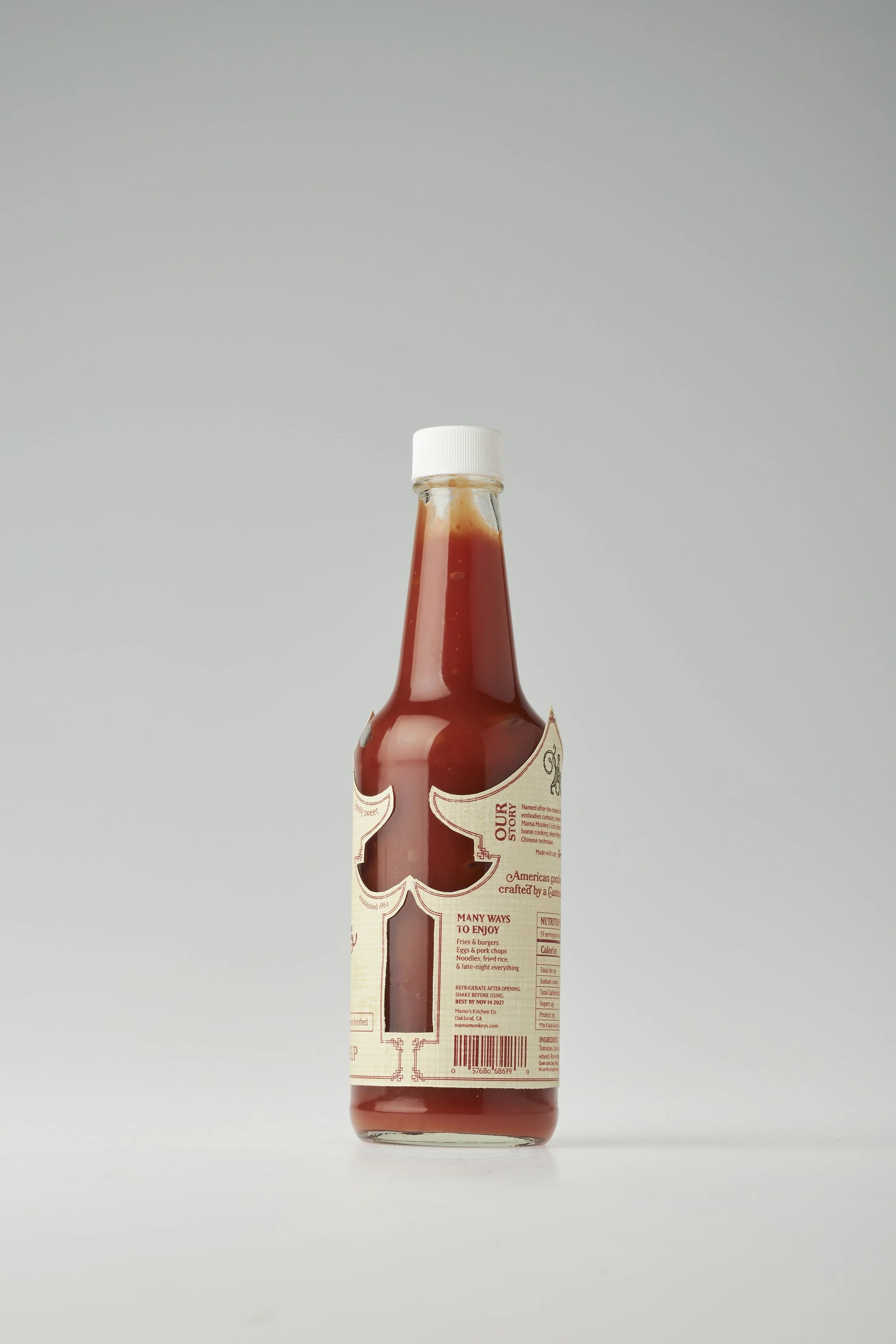 A glass bottle of ketchup with a white cap placed on a plain white surface against a light gray background.
