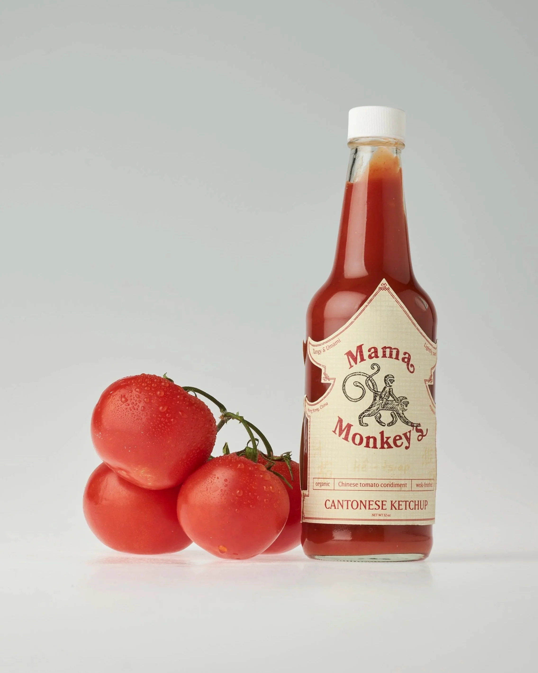 A bottle of Mama Monkey Cantonese ketchup standing next to three fresh tomatoes with water droplets on their surface.