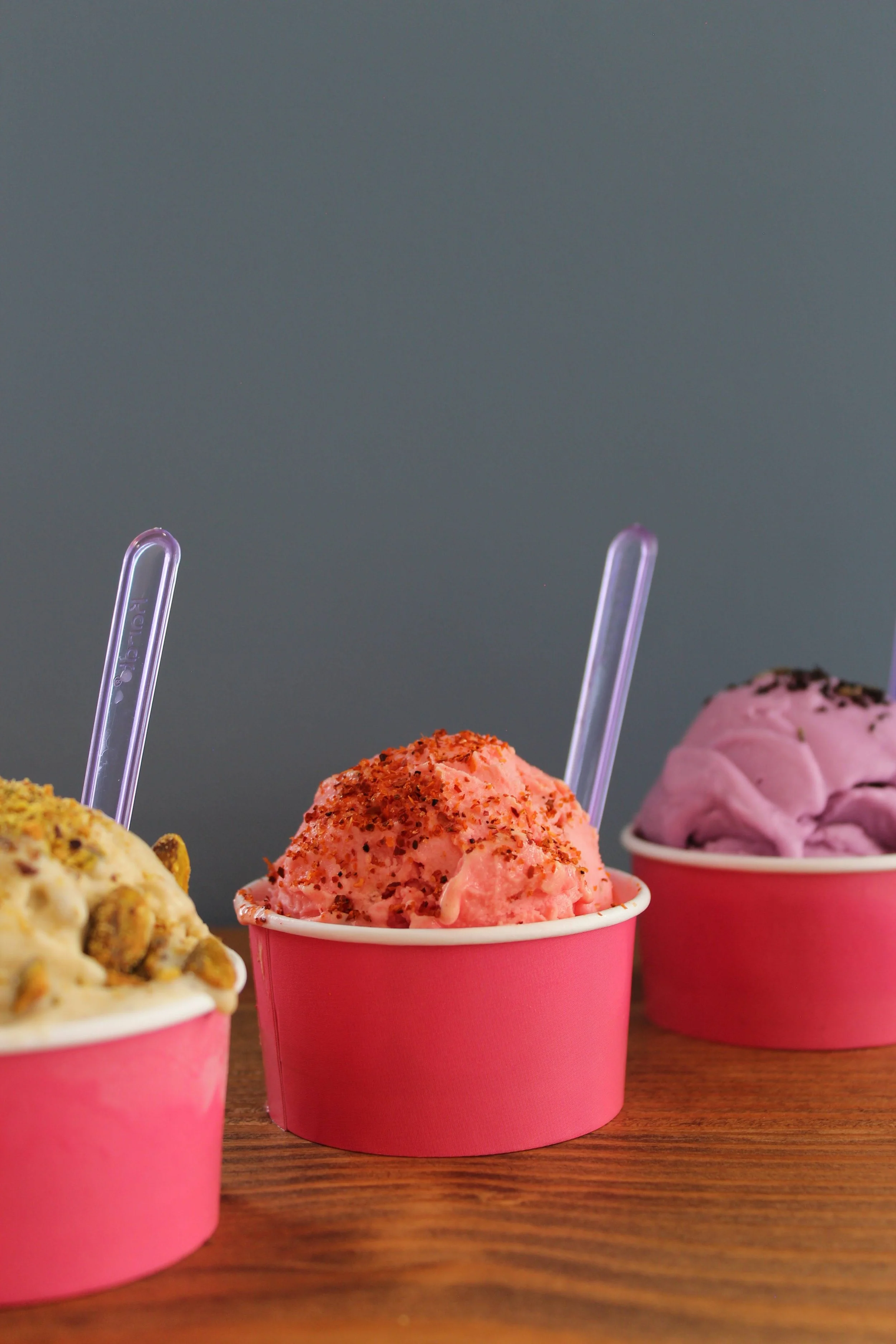 Three cups of ice cream on a wooden table, with pink cups and colorful flavors, against a plain gray background.