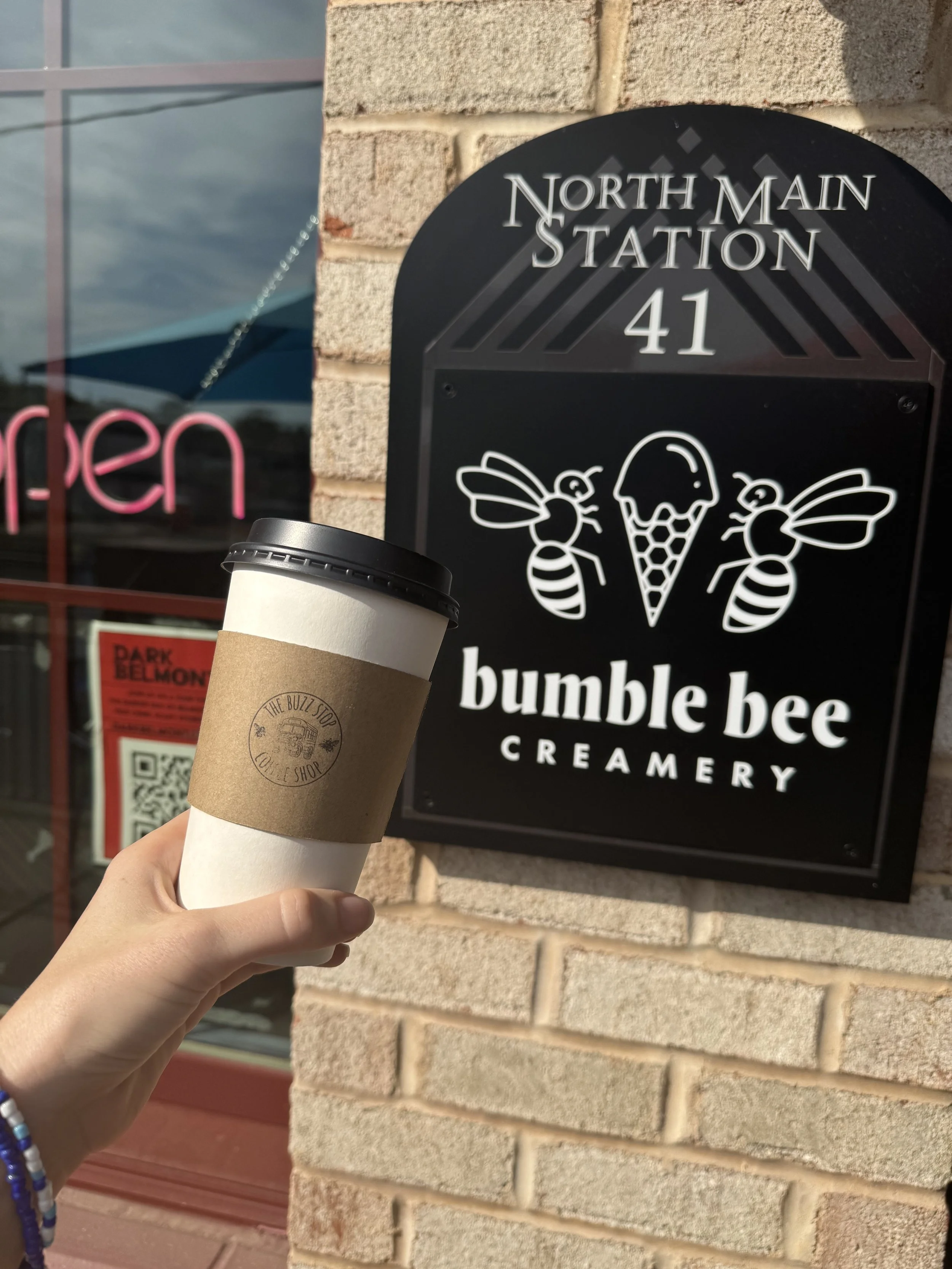 Person holding a coffee cup in front of a sign for Bumble Bee Creamery, located at North Main Station, 41. The sign features illustrations of two bees and an ice cream cone.