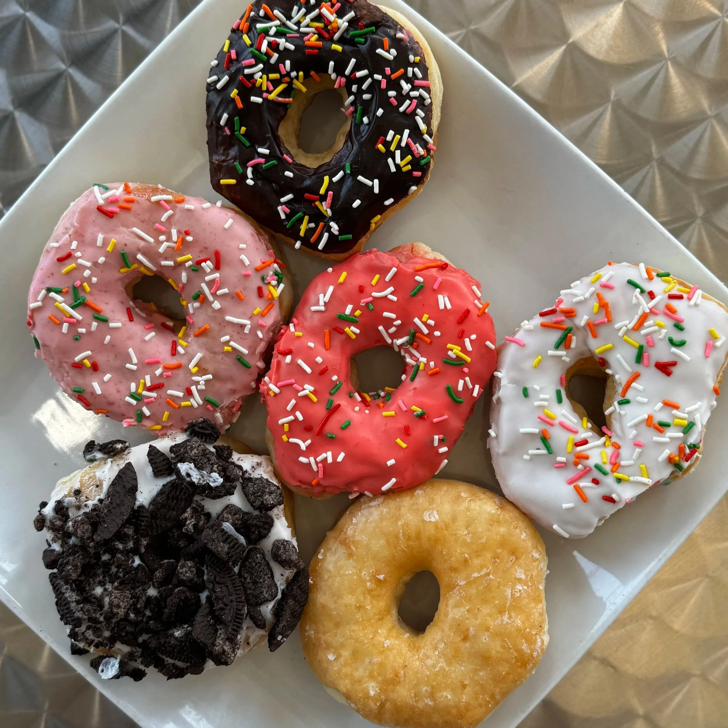 Six assorted donuts with colorful sprinkles and toppings on a white square plate. Varieties include chocolate with sprinkles, pink, red, and white frosted donuts with various toppings, and plain glazed donuts.