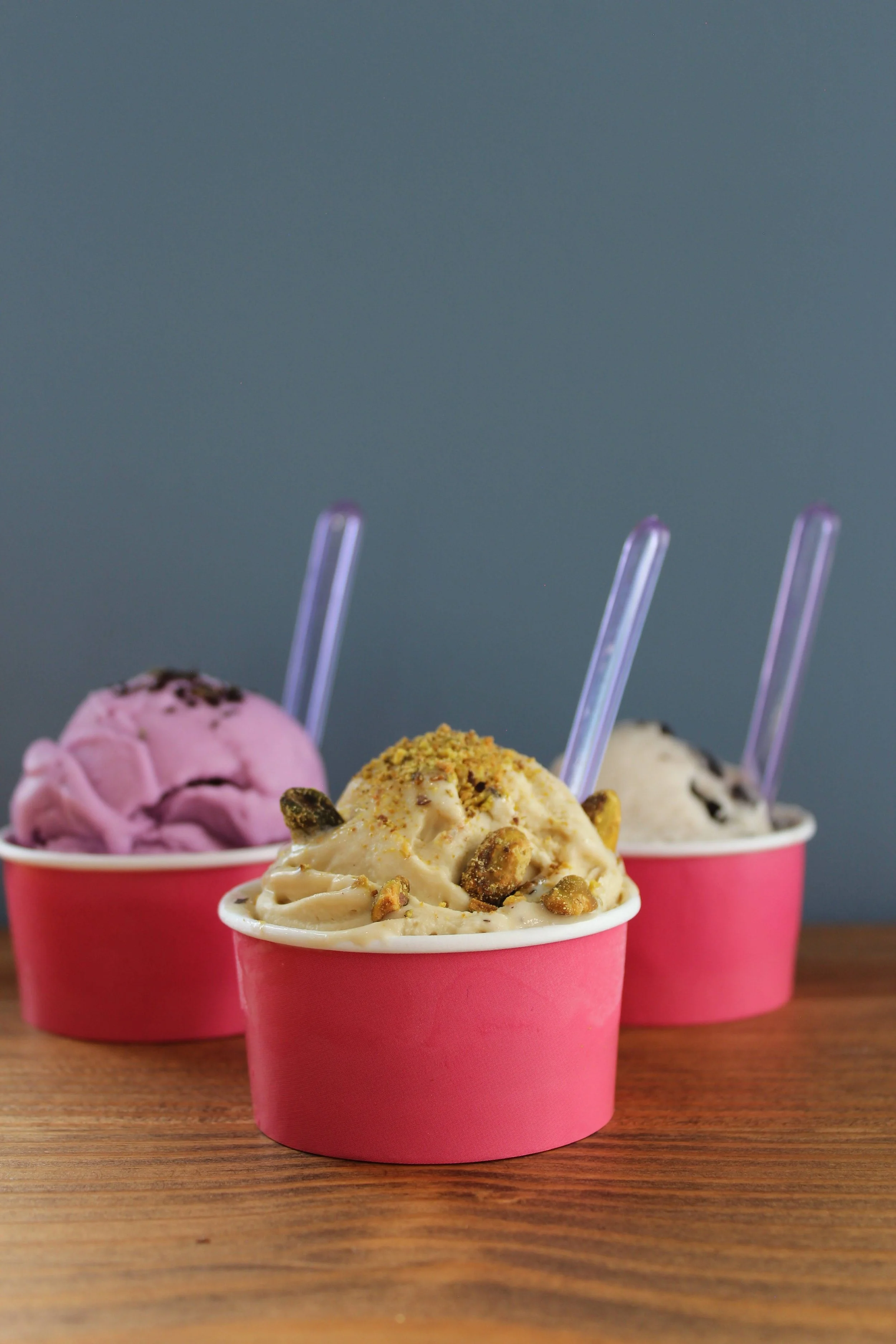 Three cups of different flavors of ice cream with clear plastic spoons on a wooden surface against a gray background.
