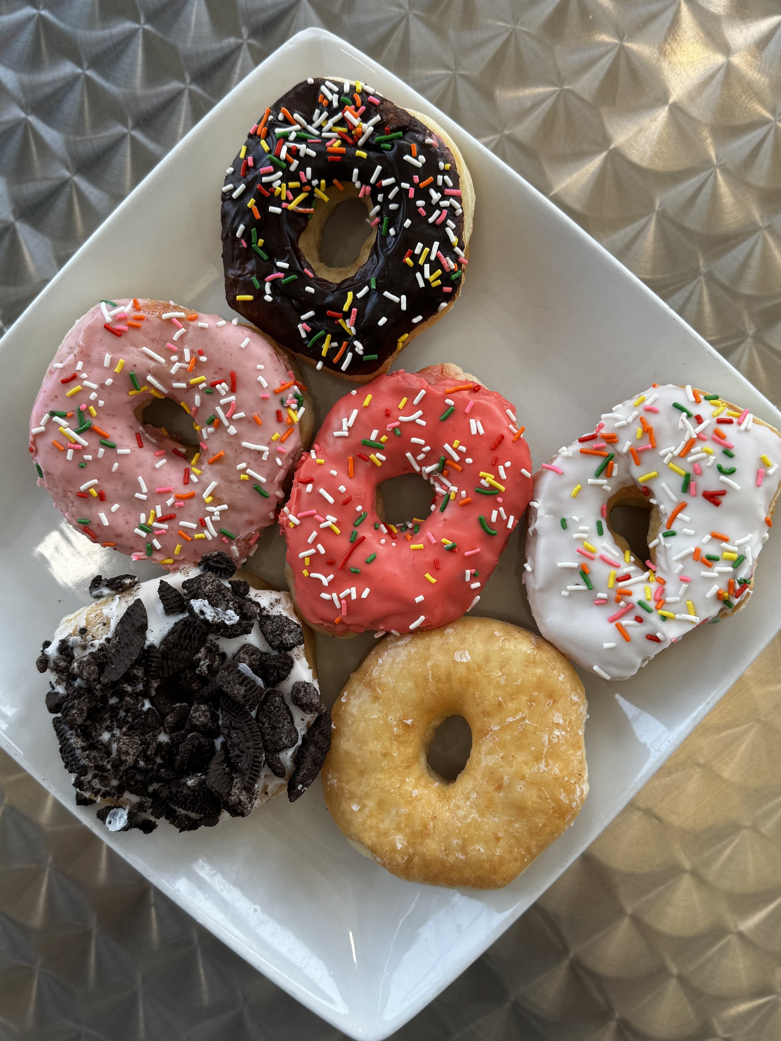 A white square plate holding six assorted donuts with colorful sprinkles and toppings, placed on a metallic textured surface. The donuts are in various flavors and toppings, including chocolate, pink, white, and vanilla.