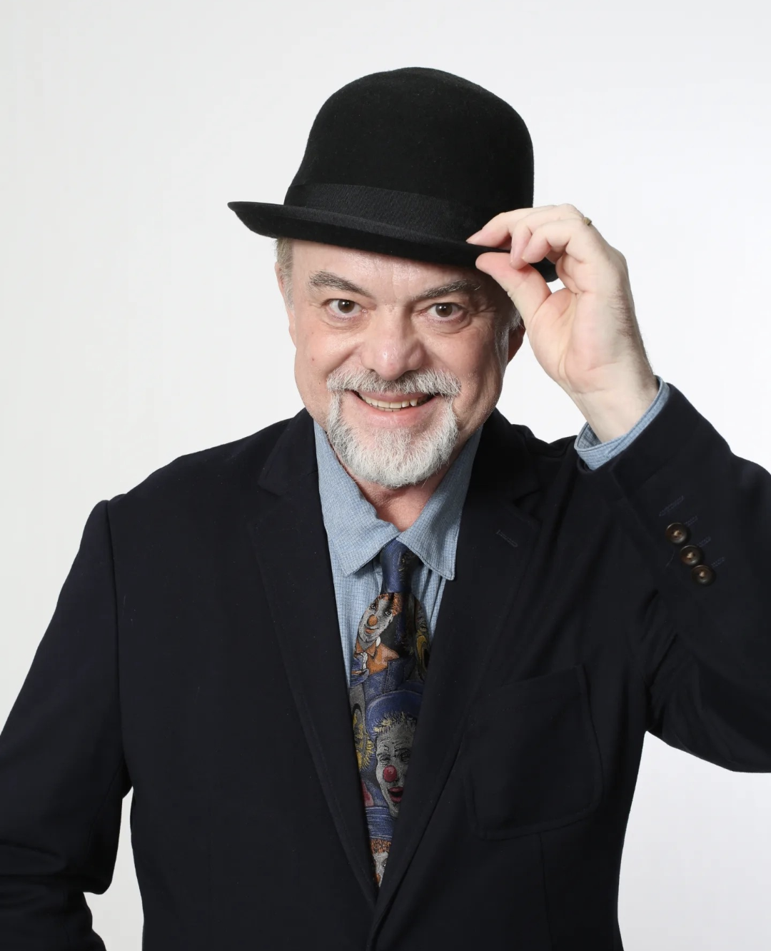 A smiling man with a gray beard and mustache wearing a black suit, a light blue shirt, a colorful tie with a clown pattern, and a black hat, adjusting his hat with one hand against a plain white background.