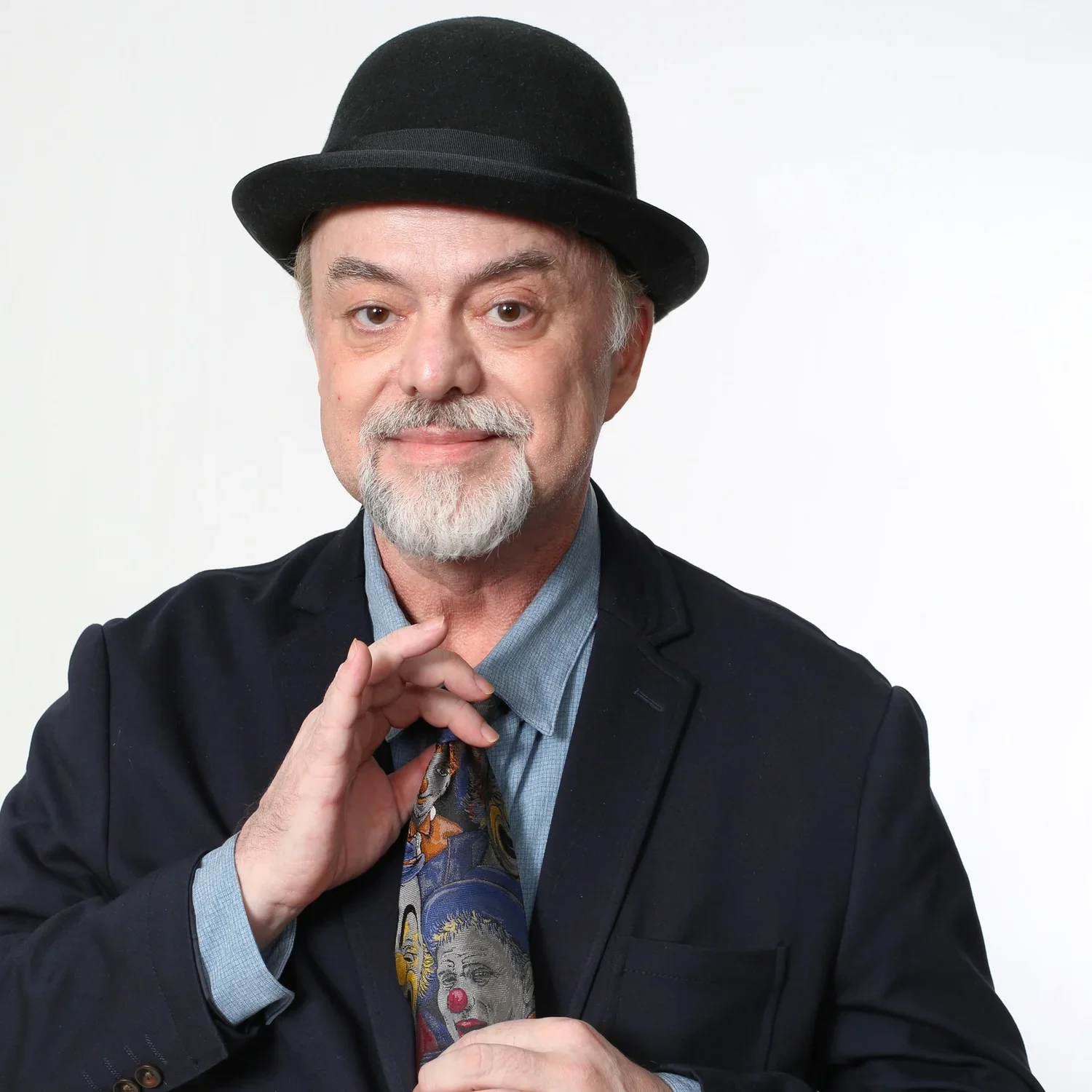 An older man with a gray beard and mustache wearing a black hat, dark blazer, light blue shirt, and patterned tie, holding his glasses with one hand, against a plain white background.