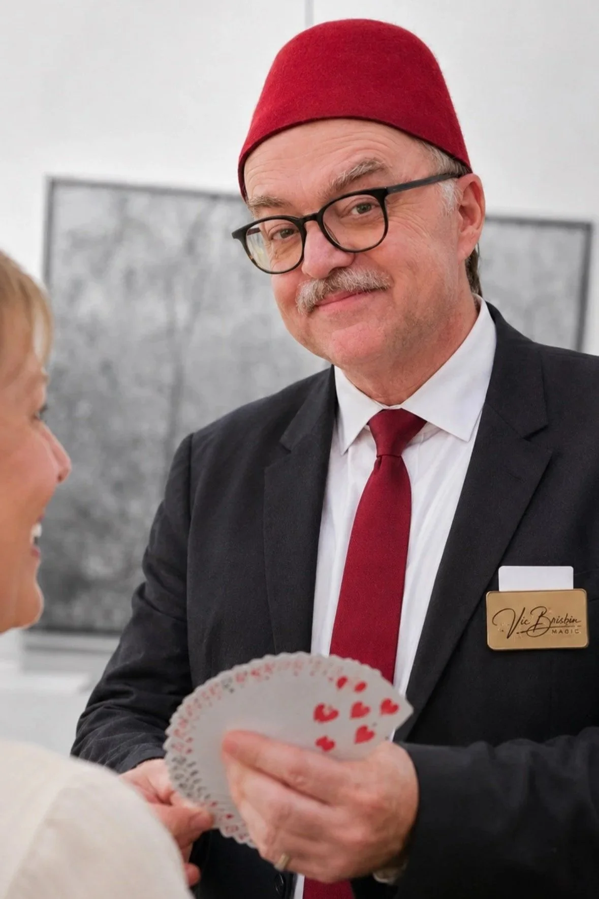 A smiling man with a gray beard and mustache wearing a black suit, a light blue shirt, a colorful tie with a clown pattern, and a black hat, adjusting his hat with one hand against a plain white background.