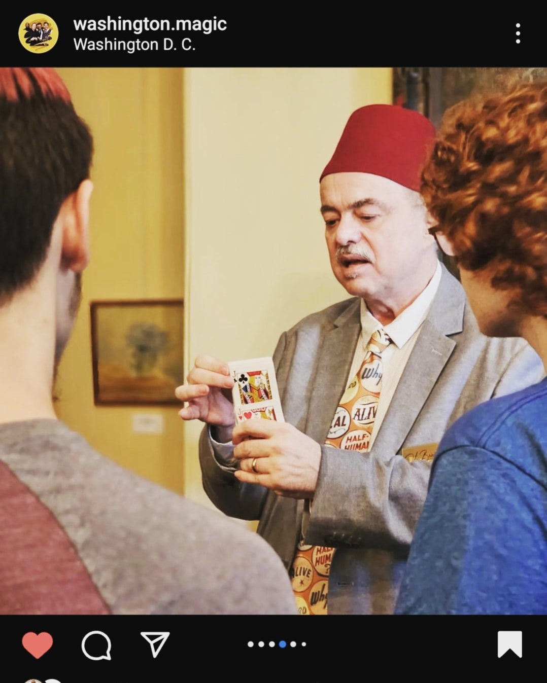 A magician in a suit and tie, wearing a red fez, demonstrating a trick with playing cards to three young men, inside a room with framed artwork on the wall.