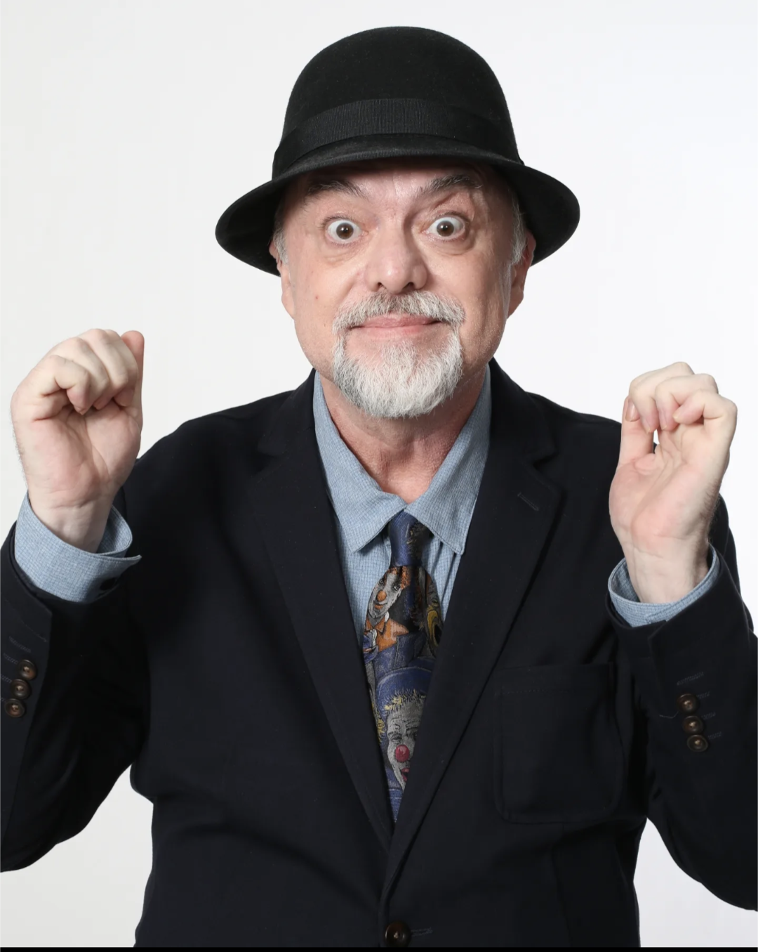 A cheerful middle-aged man with a beard and mustache, wearing a black hat, navy blazer, blue shirt, and patterned tie, smiling with fists clenched in excitement.