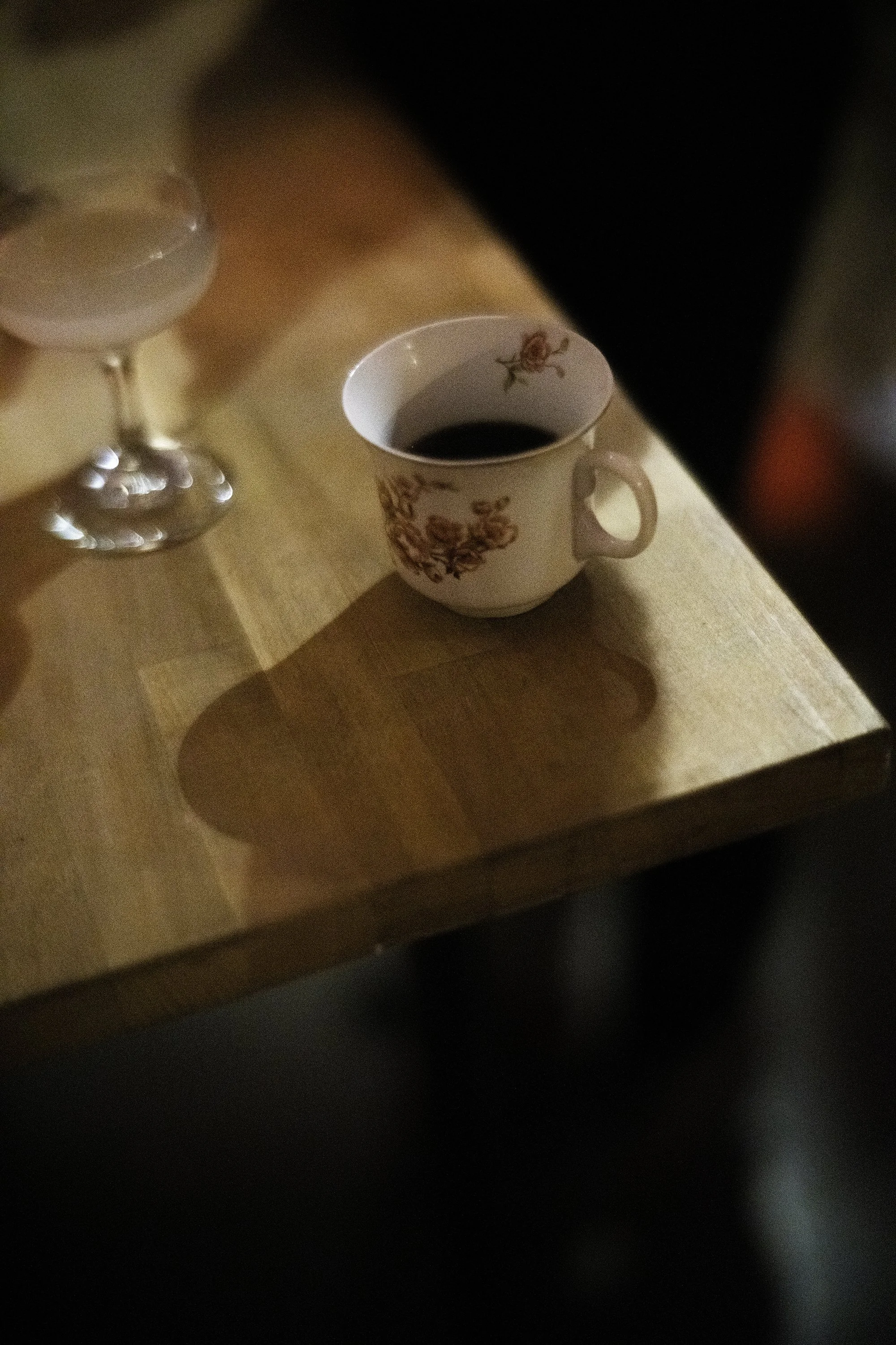 A moody photo of an in-focus teacup and glass of wine sitting half full on the corner of a wooden table.