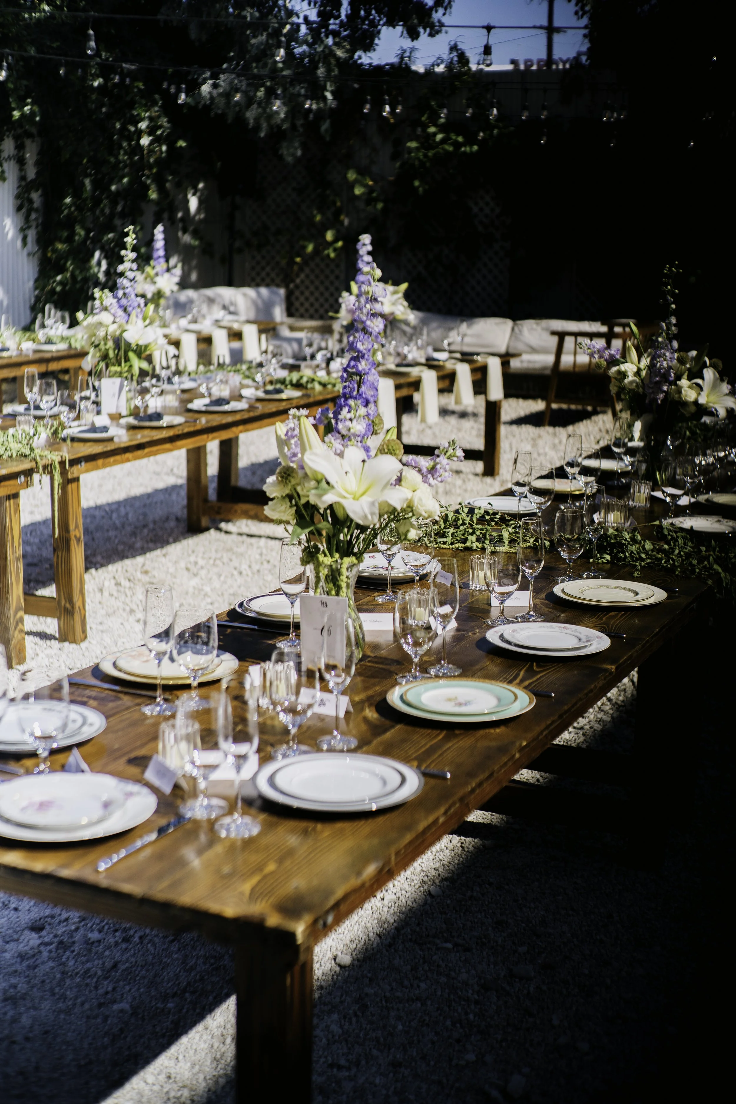A beautiful sunlit afternoon showing three farm tables set up for 10 or more people for a wedding event. Each dinner and salad plate setting is different, uniquely mixed and matched together for thematically curated to look like a part of collection.