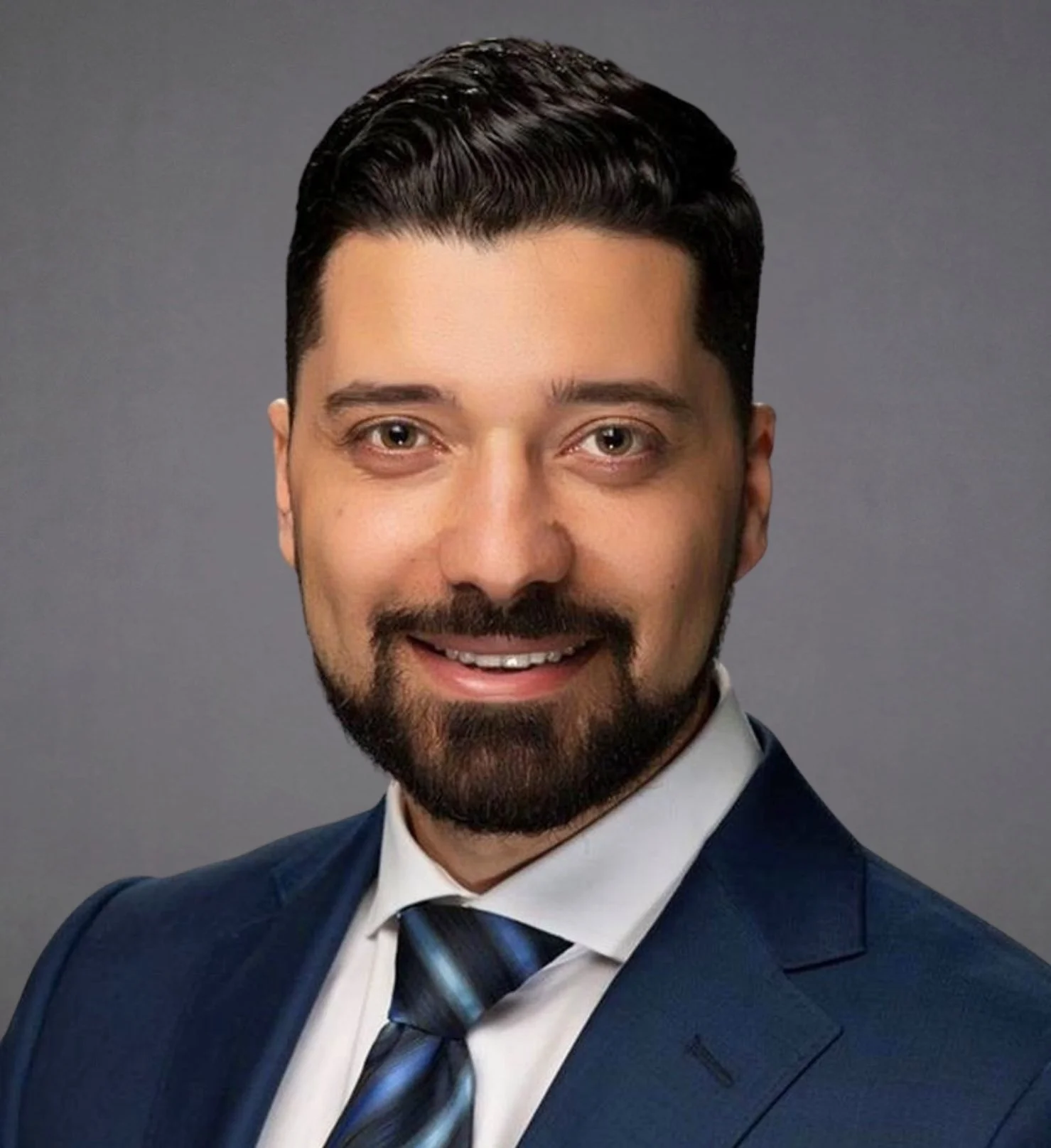 Professional headshot of a man with dark hair, beard, wearing a dark suit, white shirt, and striped tie, smiling against a gray background.