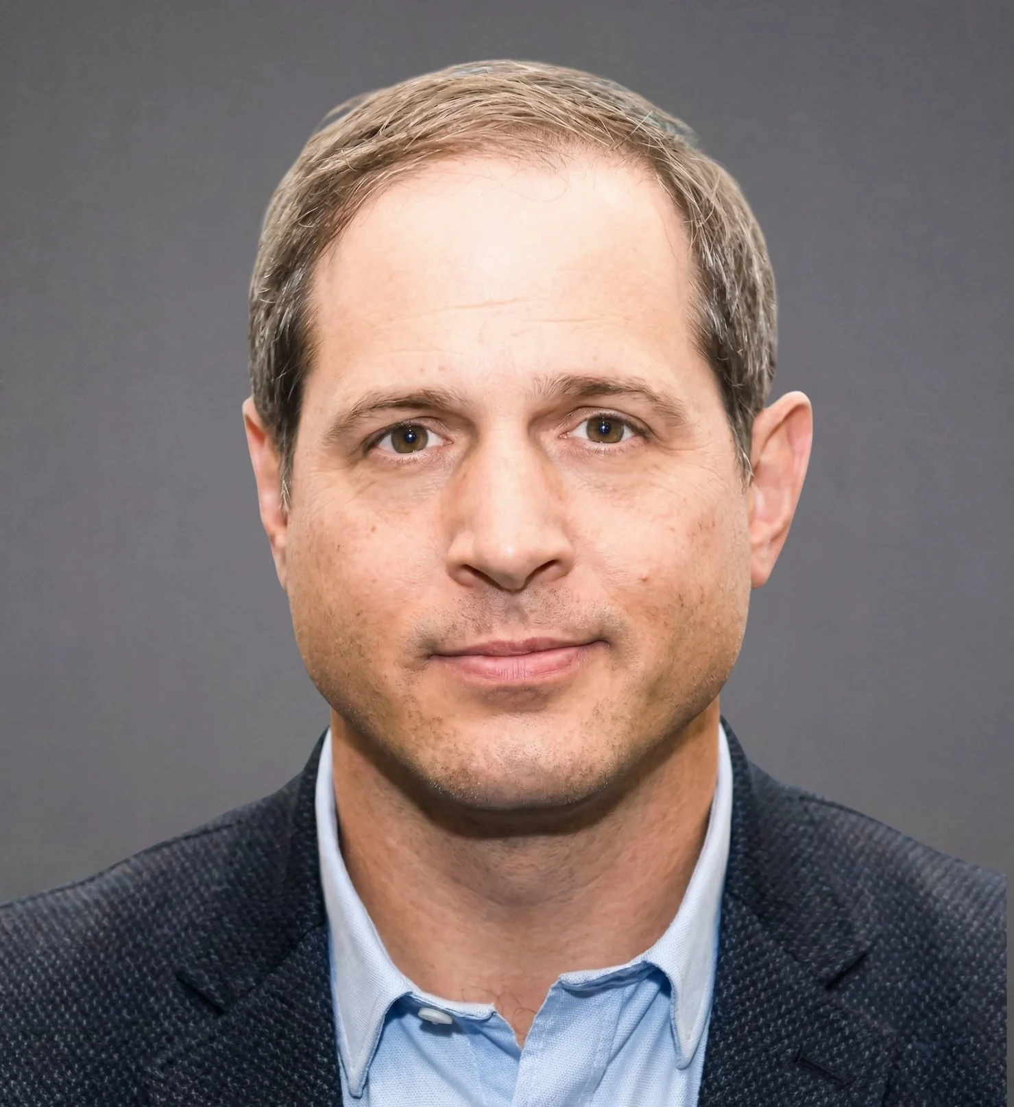 A portrait of a man with short brown hair, wearing a blue shirt and a dark blazer, against a neutral gray background.