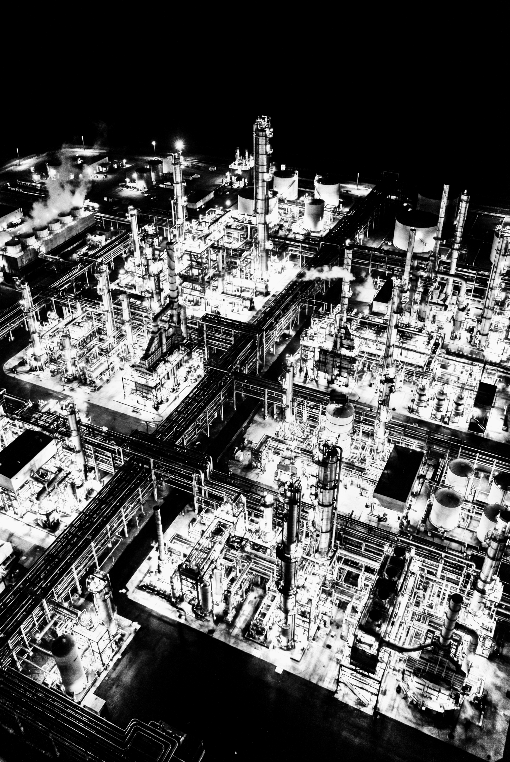 Black and white aerial view of an industrial refinery at night with multiple towers, pipes, and smokestacks emitting steam or smoke.