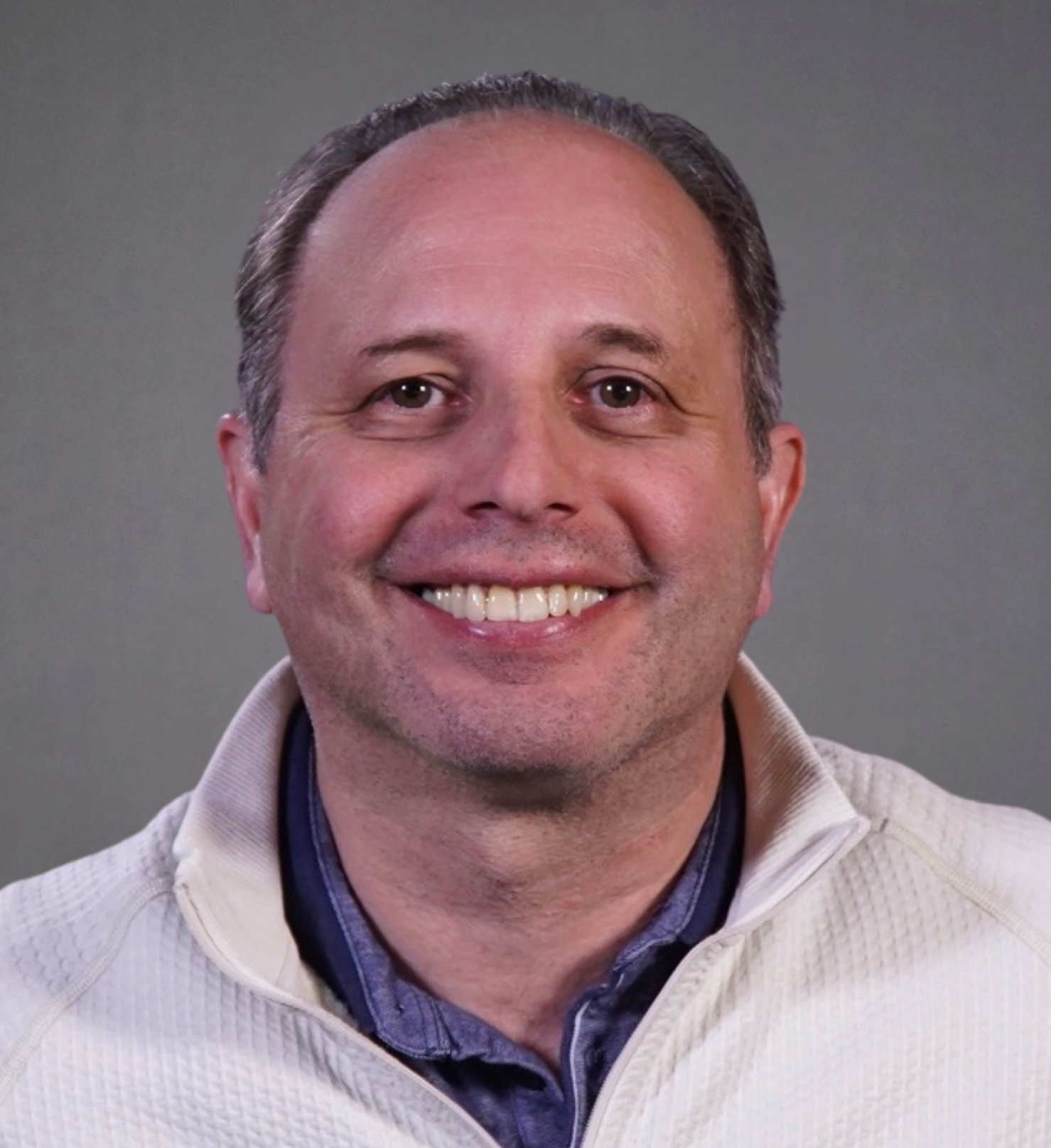 Headshot of a smiling middle-aged man with short brown hair, wearing a light-colored jacket over a dark shirt.