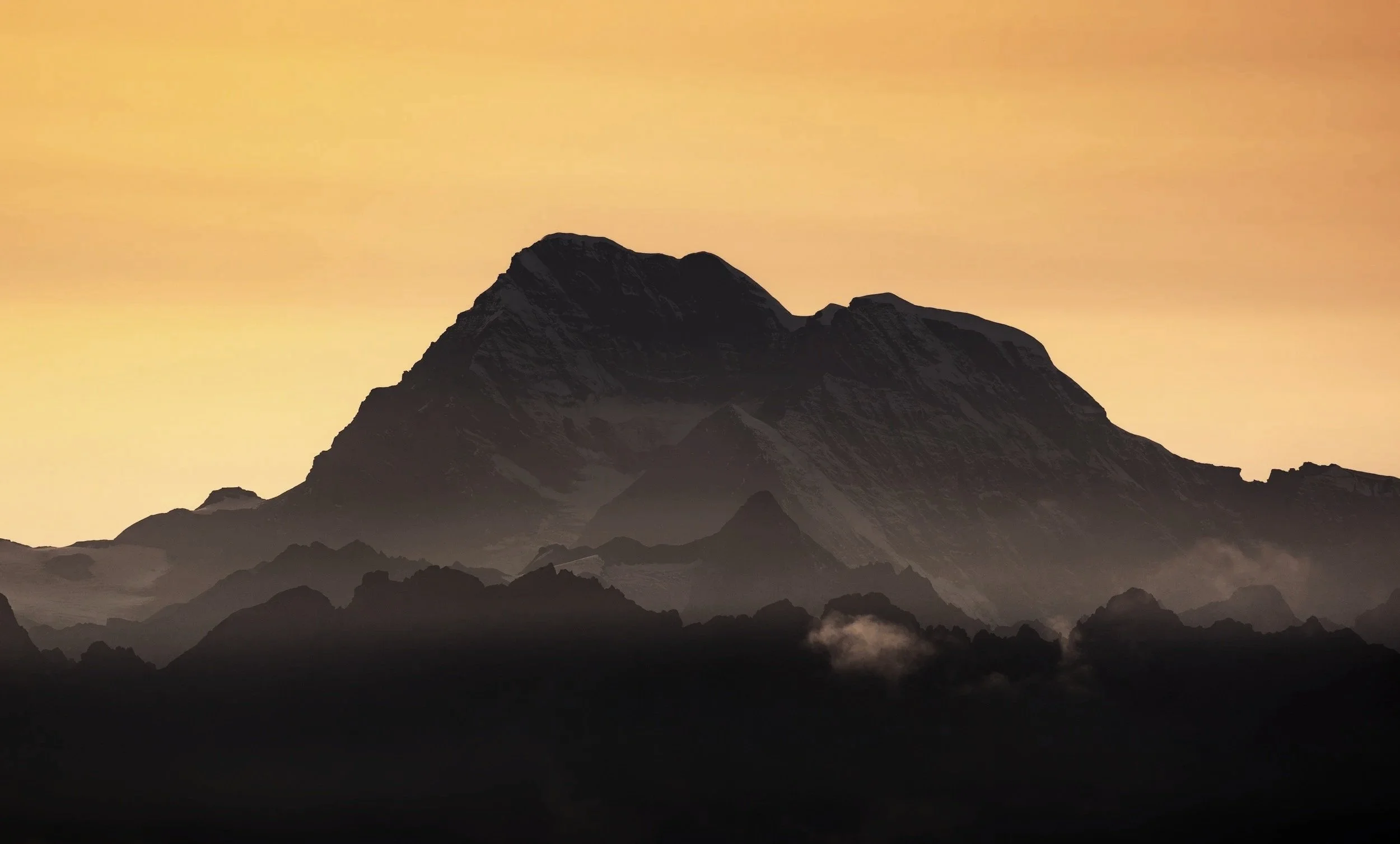 Silhouette of a mountain range at sunset with a gradient orange sky and dark clouds at the base of the mountains.