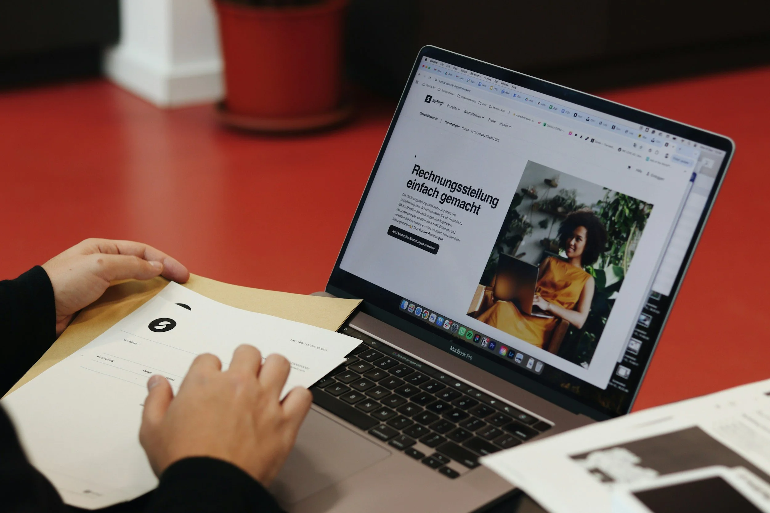 A person sitting at a red desk using a laptop with a spreadsheet and a website open, while holding a paper form.