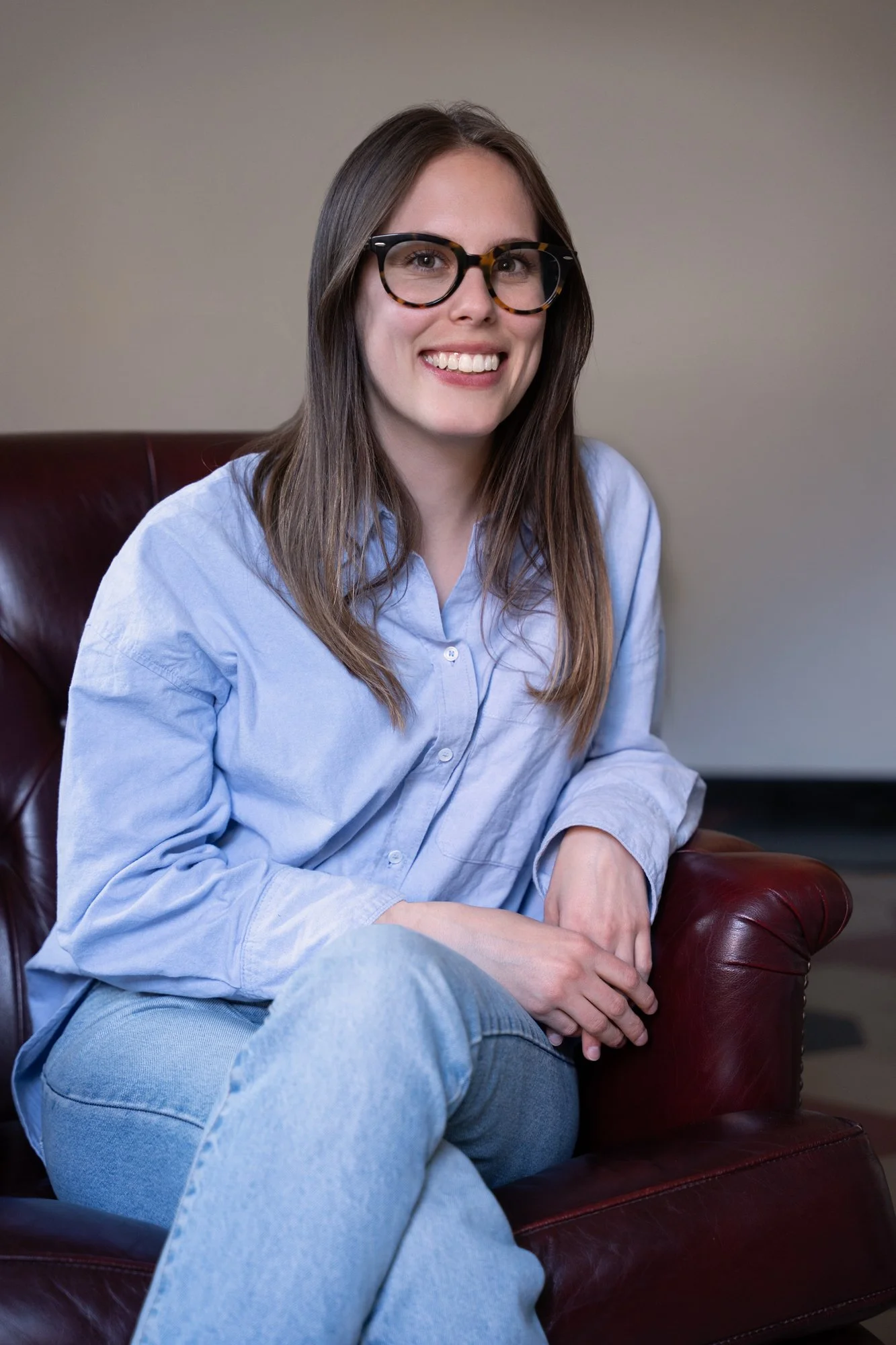A young woman with long brown hair, wearing glasses and a light blue shirt, is sitting on a dark red leather chair, smiling at the camera.