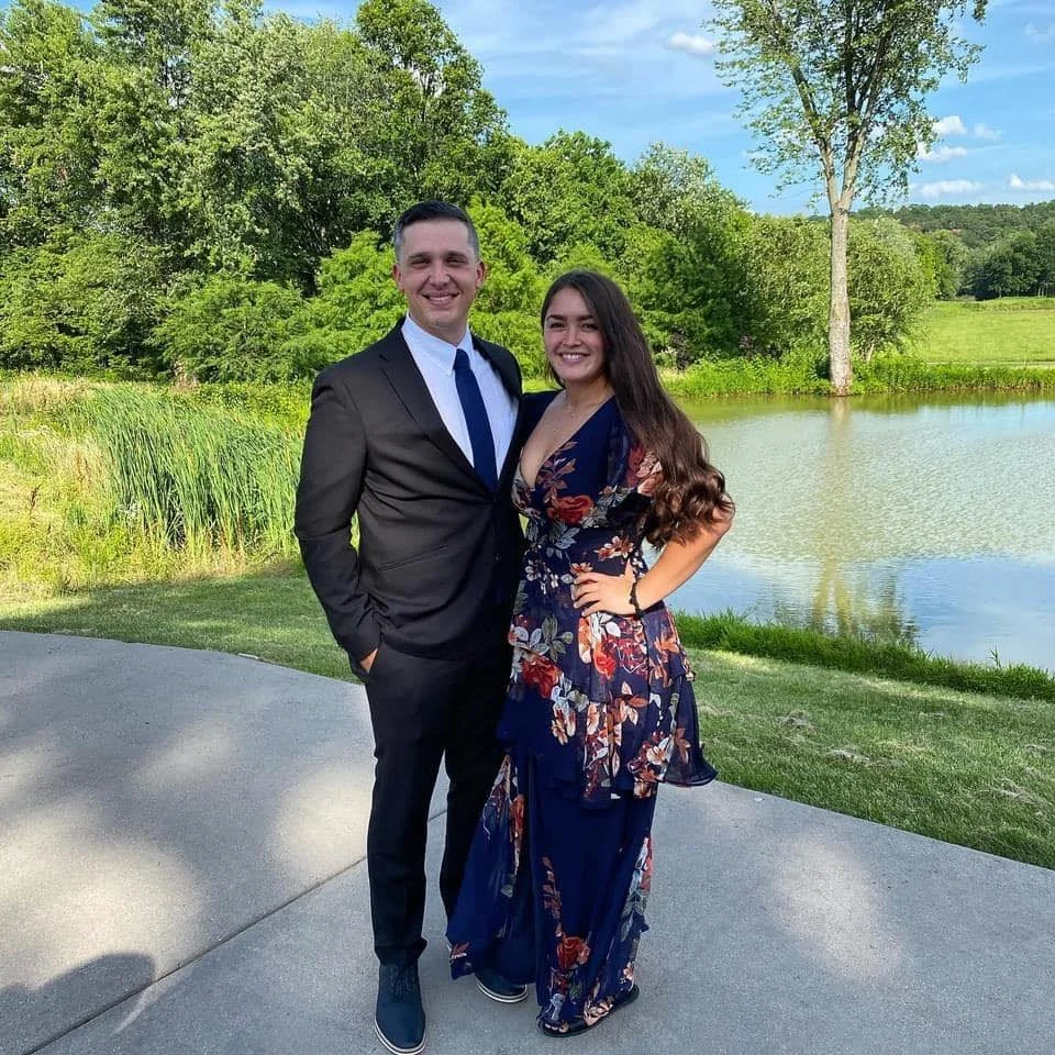 A smiling man in a suit and a woman in a floral dress standing outdoors near a pond with green trees and grass in the background.