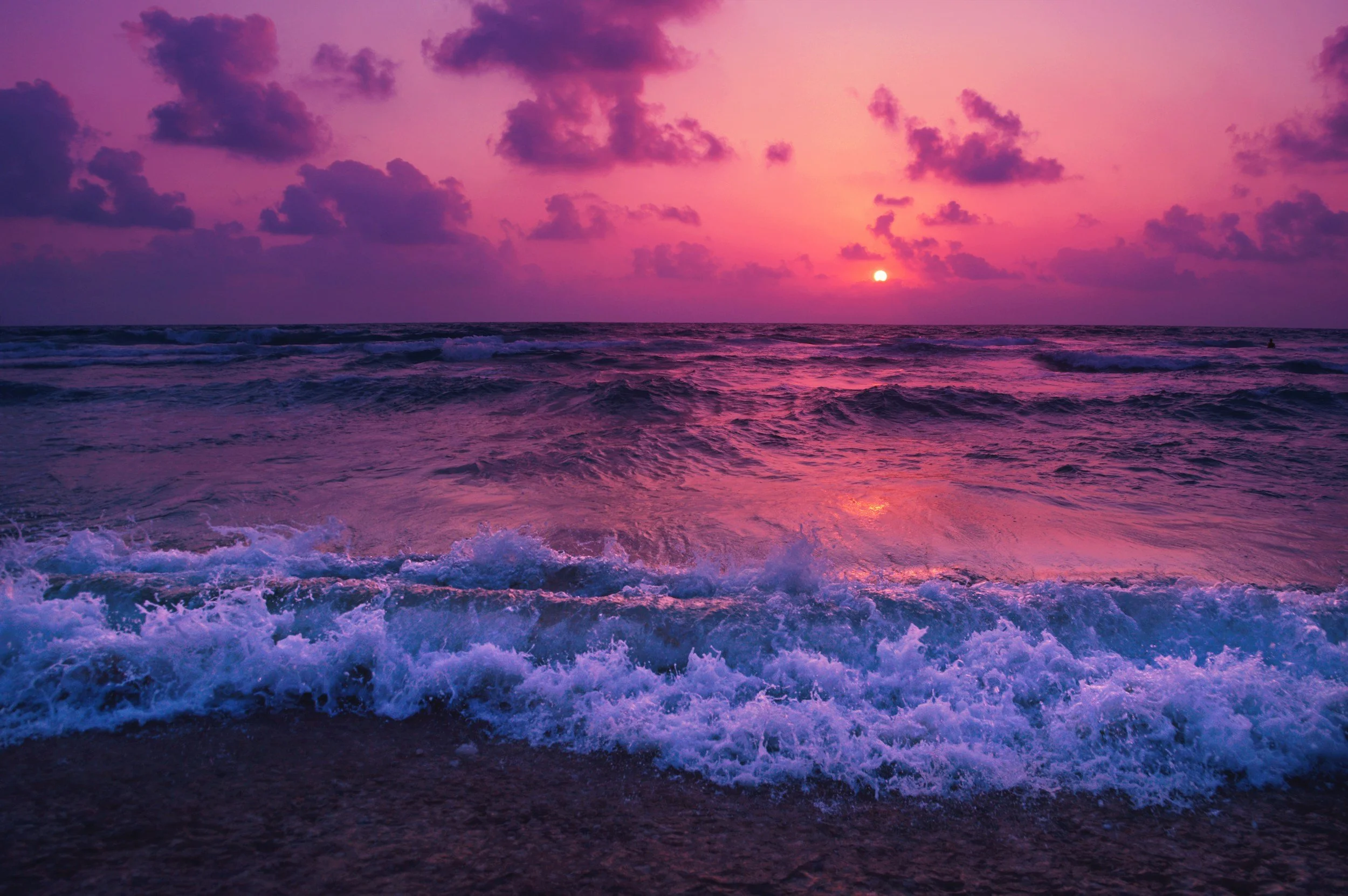 Sun setting over the ocean with pink and purple clouds in the sky, reflecting on the water and waves crashing onto the shore.