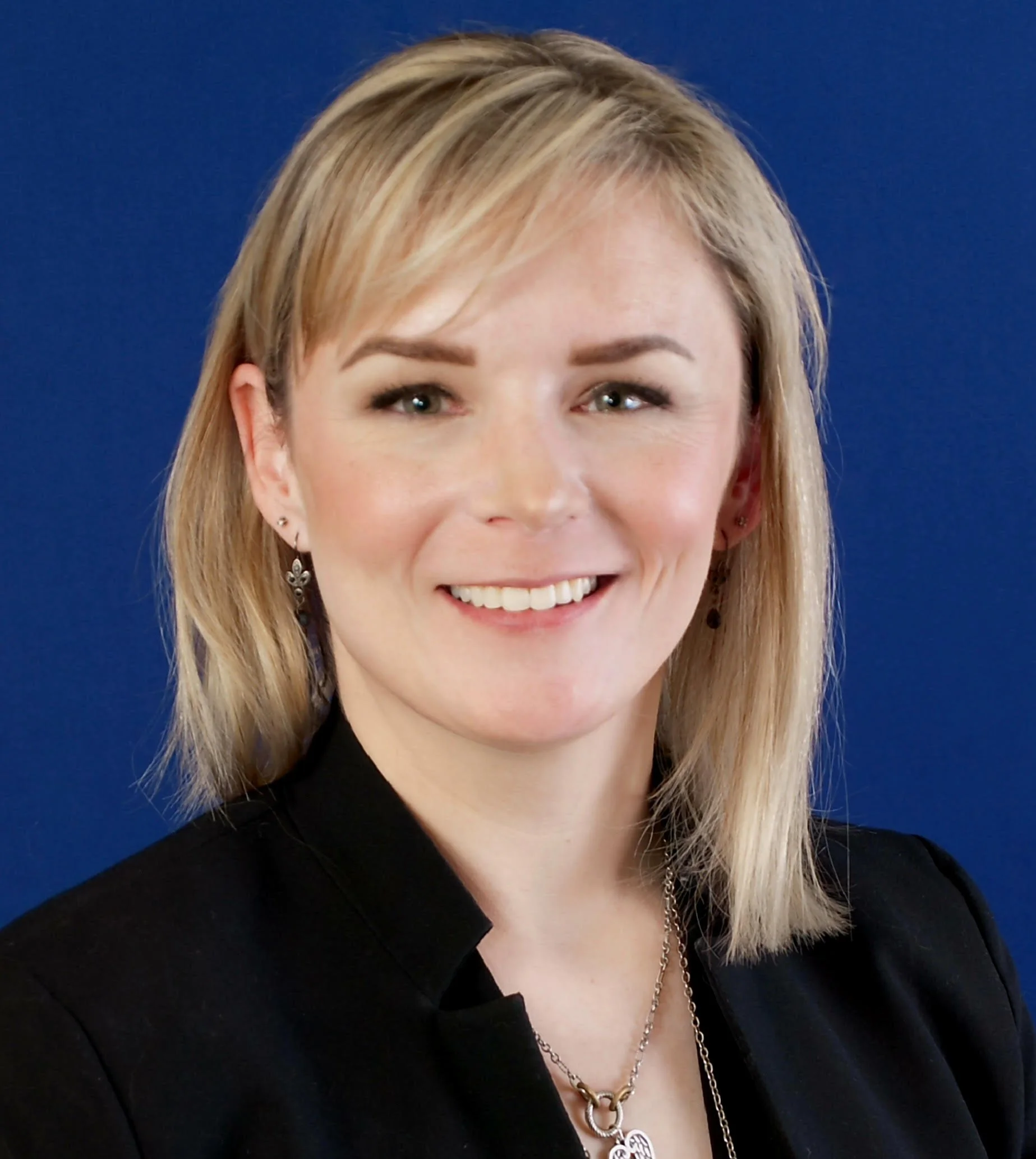 A woman with shoulder-length blonde hair smiling, wearing earrings, a black top, and layered necklaces against a dark blue background.