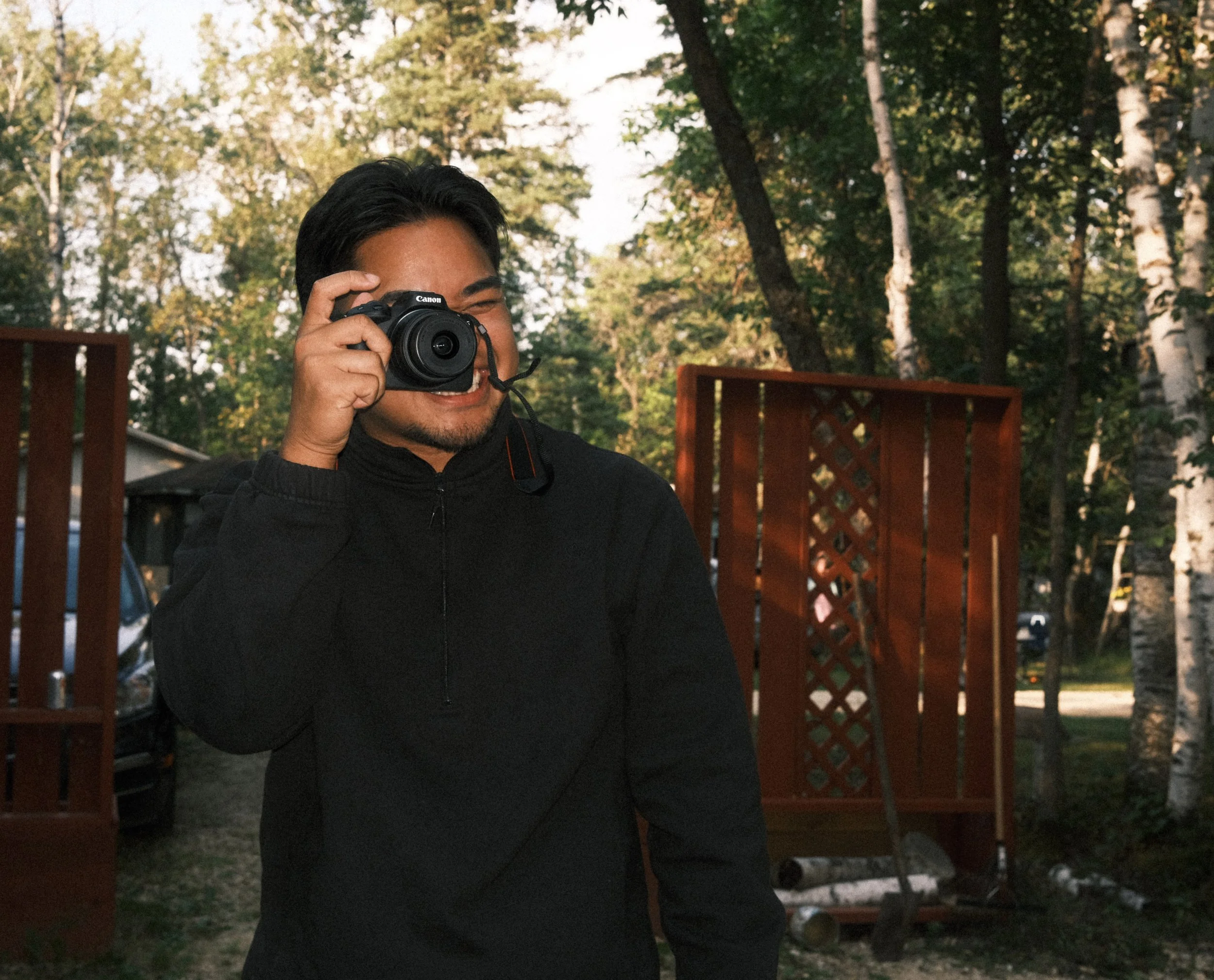 A man smiling while holding a camera up to his eye, standing outdoors with trees and wooden structures in the background.