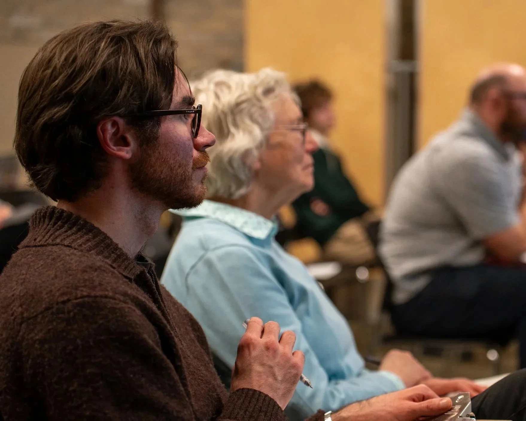 Side view of several people seated and attending a presentation or lecture, focusing on a young man with glasses and a brown sweater, an older woman with white hair and glasses, and a man in a light-colored shirt, all paying attention.