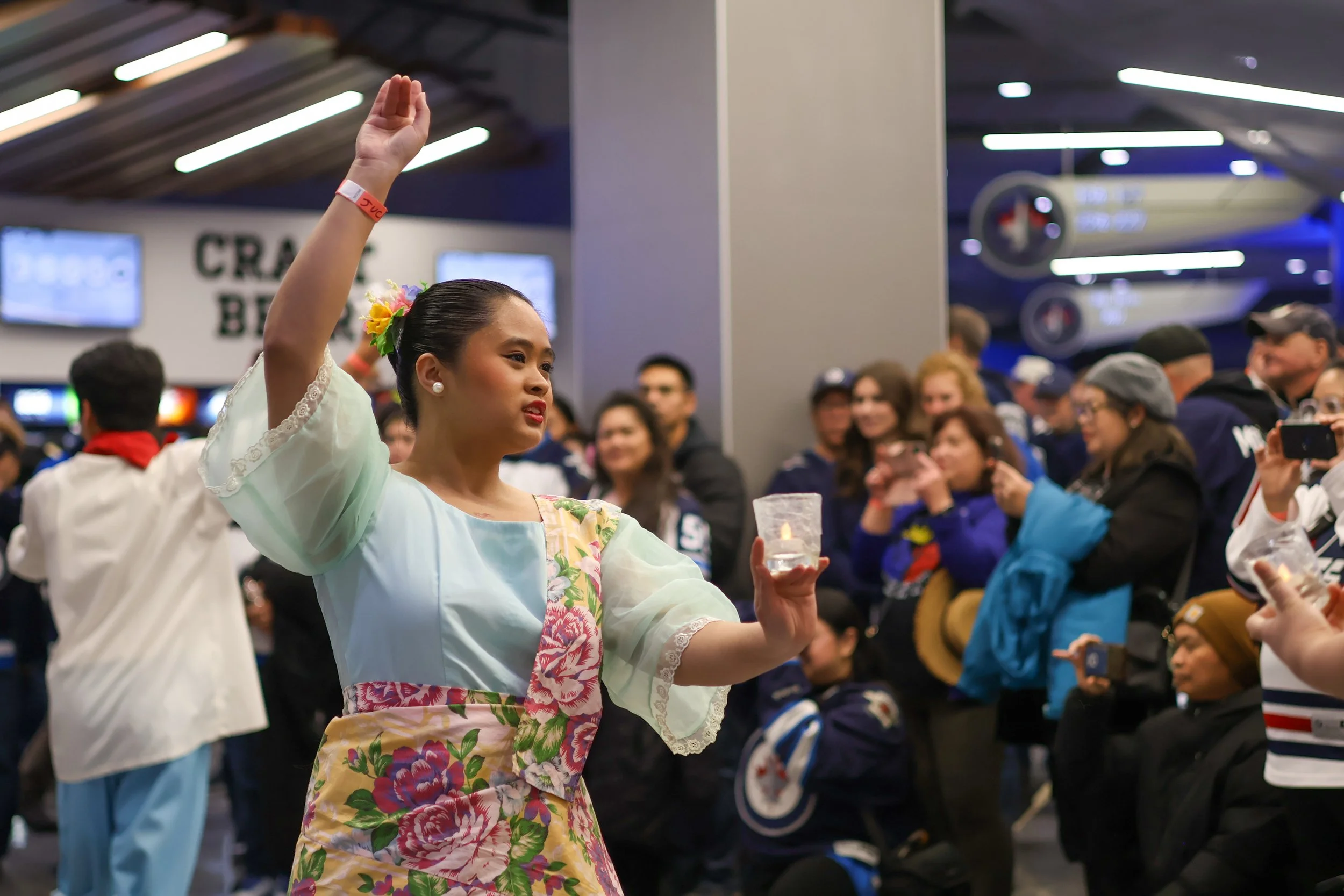 A woman in traditional clothing holding a candle and performing a cultural dance at an indoor event surrounded by a crowd of onlookers taking photos.