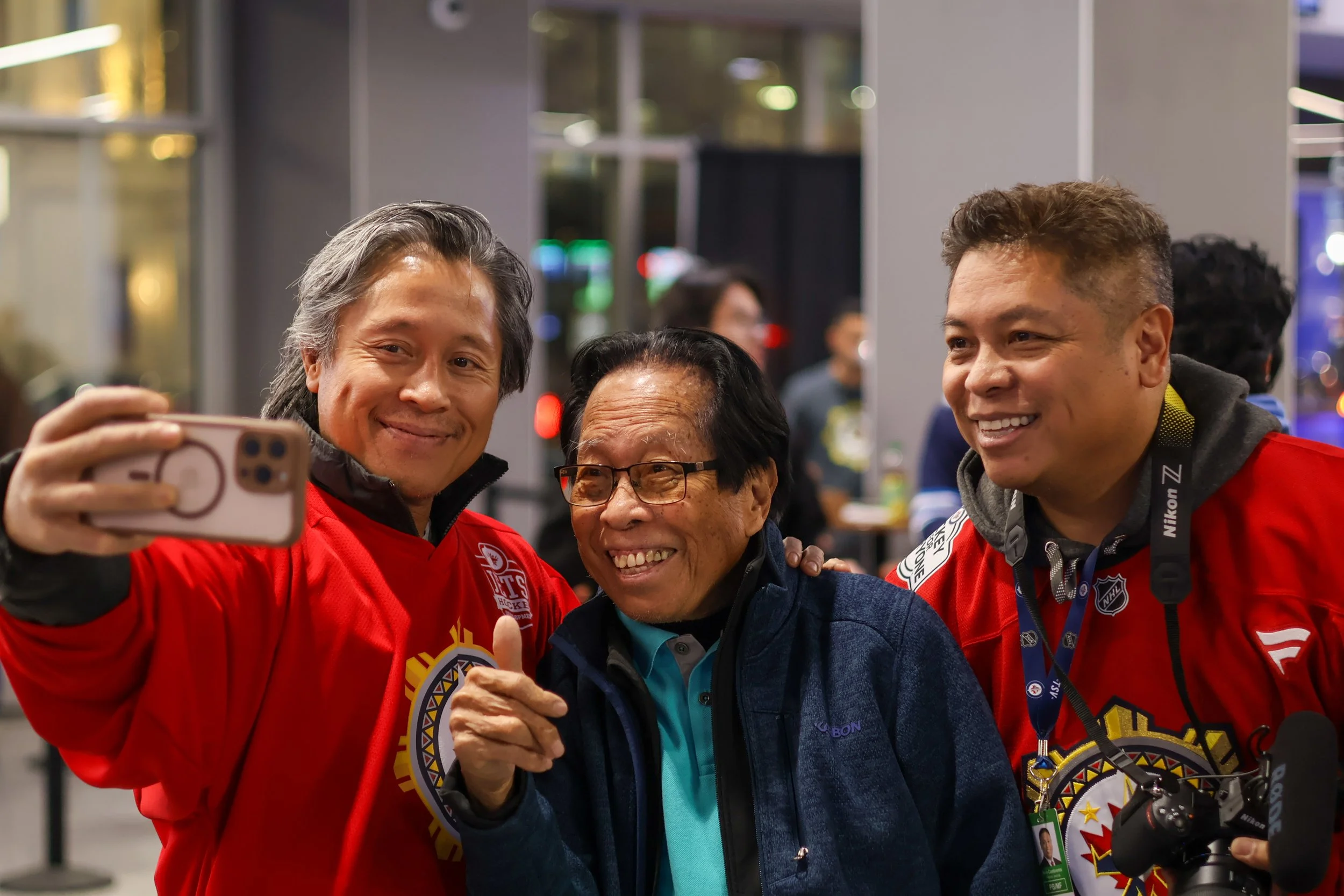 Three men smiling and taking a selfie together in an indoor setting. The two men on the sides are wearing red hockey jerseys, and the man in the middle is wearing glasses and a dark jacket.