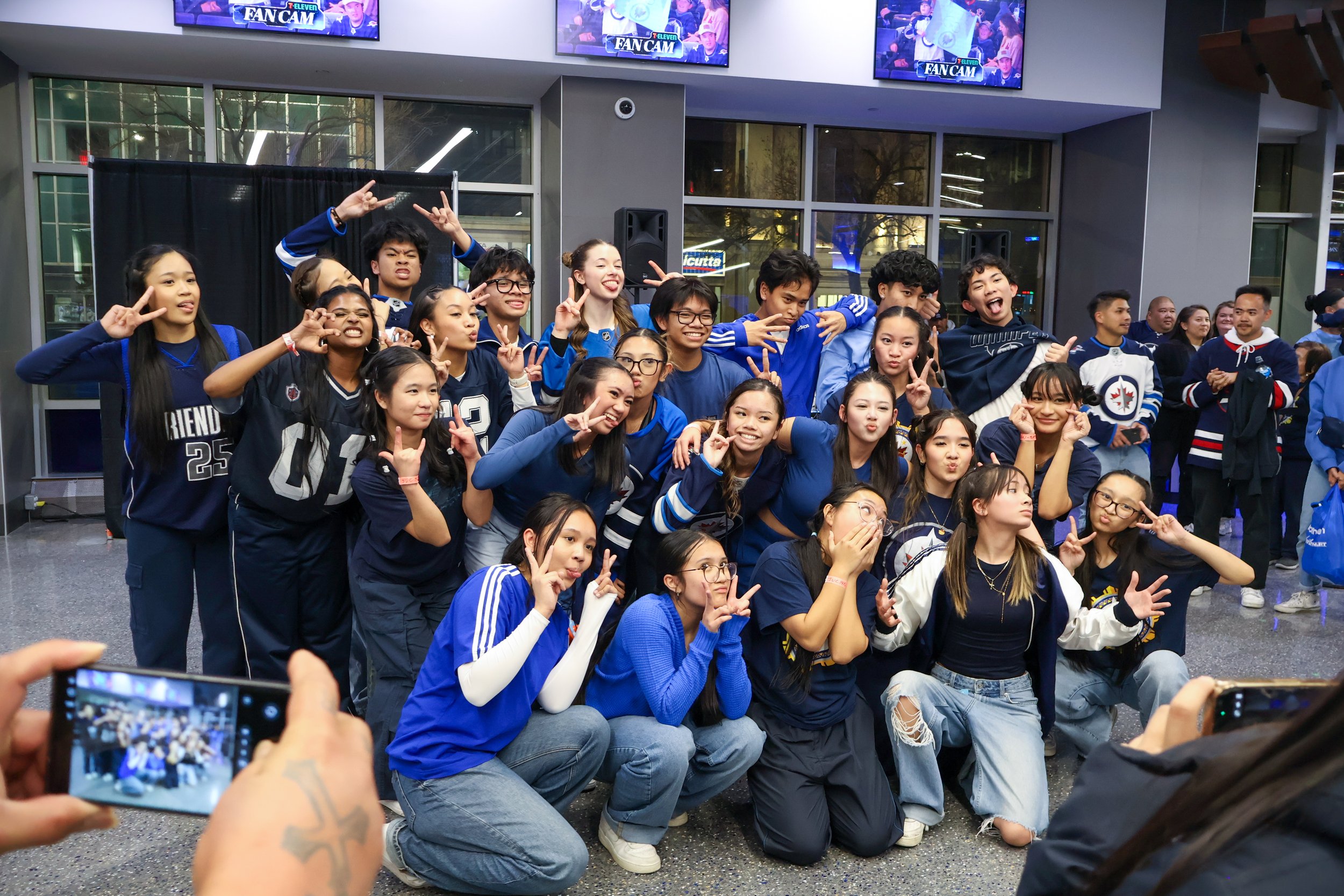 Group of young people, mostly girls, posing for a photo at a sporting event wearing blue and white jerseys and apparel, with some making peace signs and playful gestures. Others are standing or crouching in front and behind, in a modern indoor setting with multiple screens displaying the 'FAN CAM' logo.
