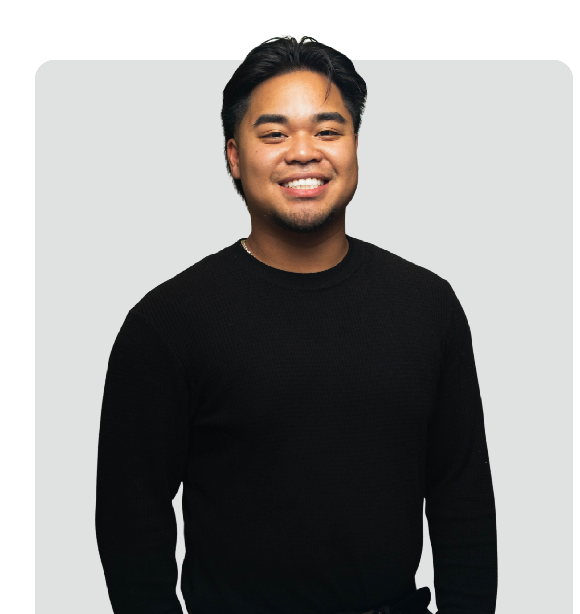 A young man with dark hair, smiling, wearing a black sweater, against a light gray background.