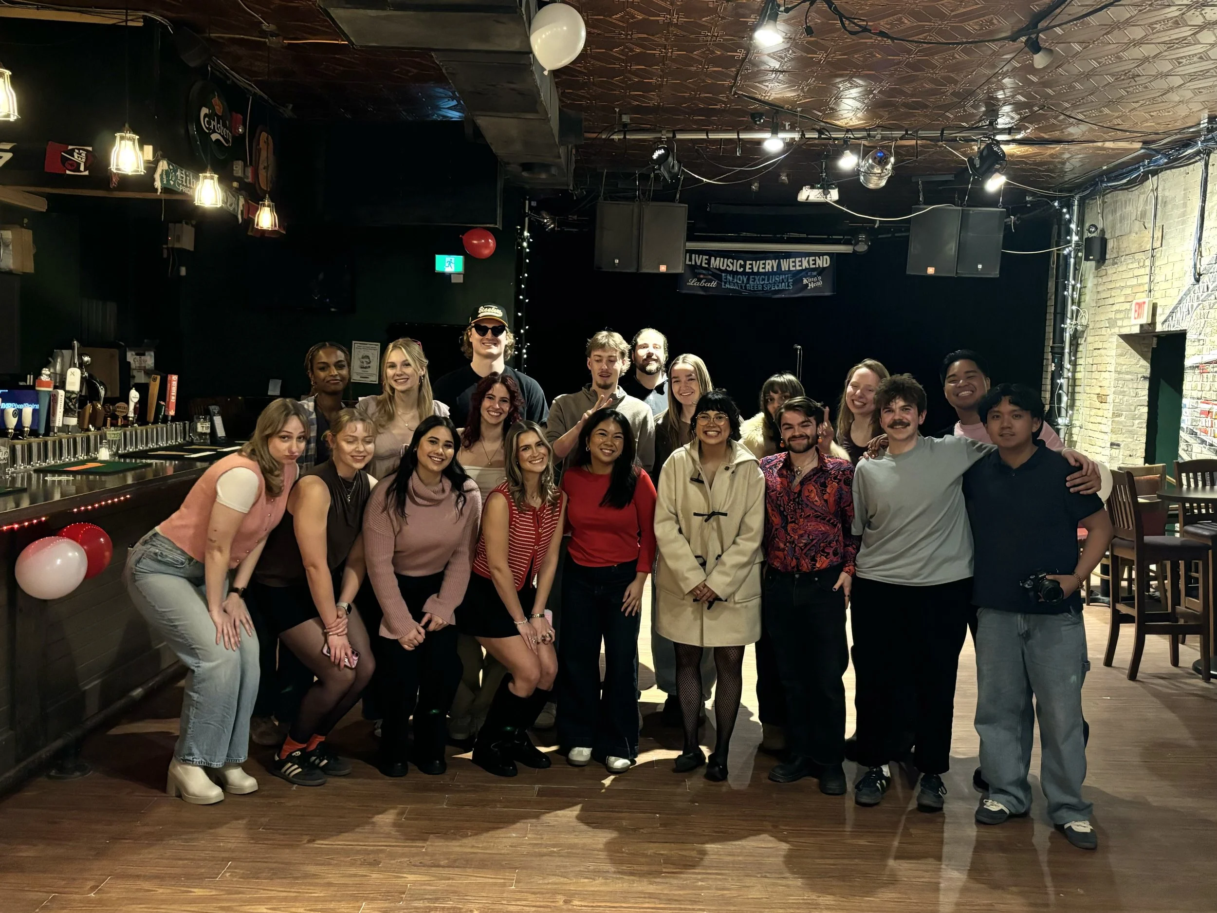 A group of 17 young adults posing in a bar or music venue with a stage, bar area on the left, and brick wall on the right. They are smiling, standing close together, and dressed casually.