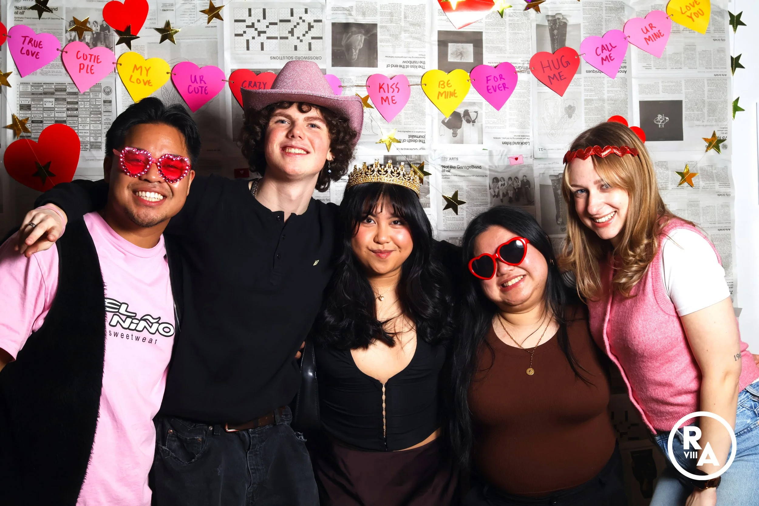 Group of five young adults celebrating Valentine's Day with props and decorations, standing in front of a backdrop decorated with heart-shaped notes and gold stars.