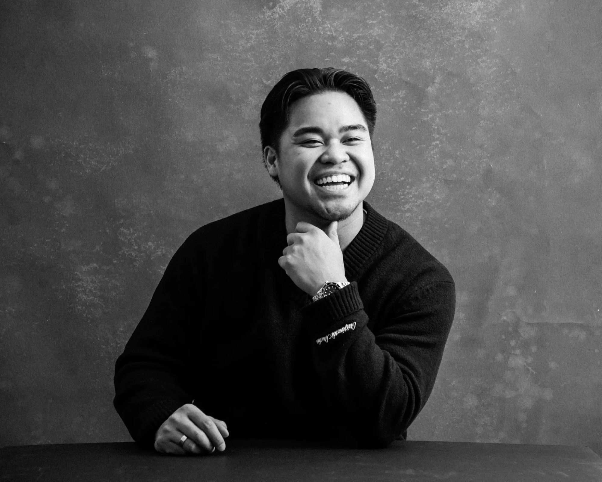 Black and white portrait of a smiling young man with dark hair, wearing a dark sweater, sitting at a table with his left arm resting on it, and his right hand touching his chin, against a textured background.
