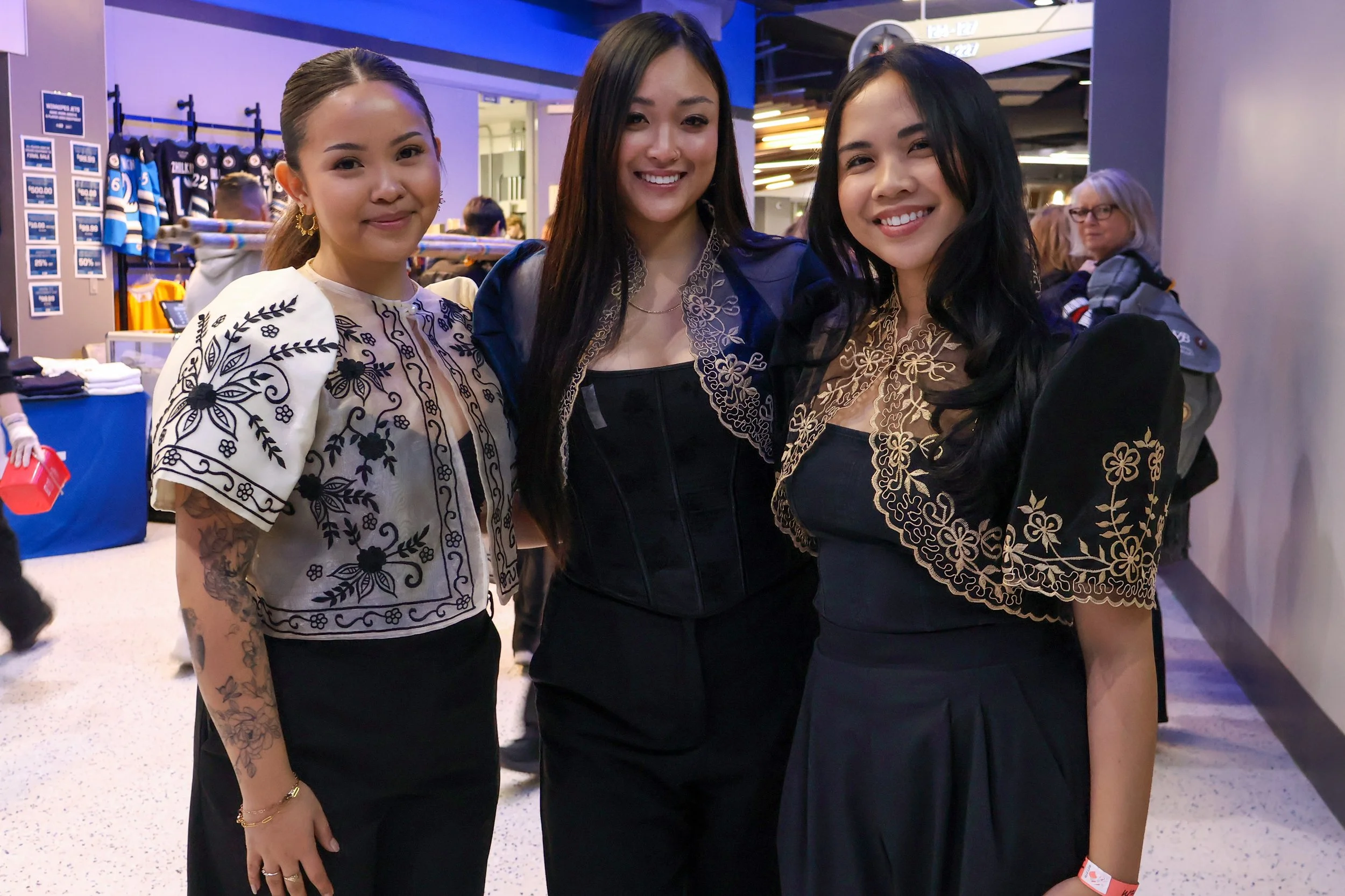 Three women standing together at an indoor event, smiling at the camera, with a merchandise booth in the background.