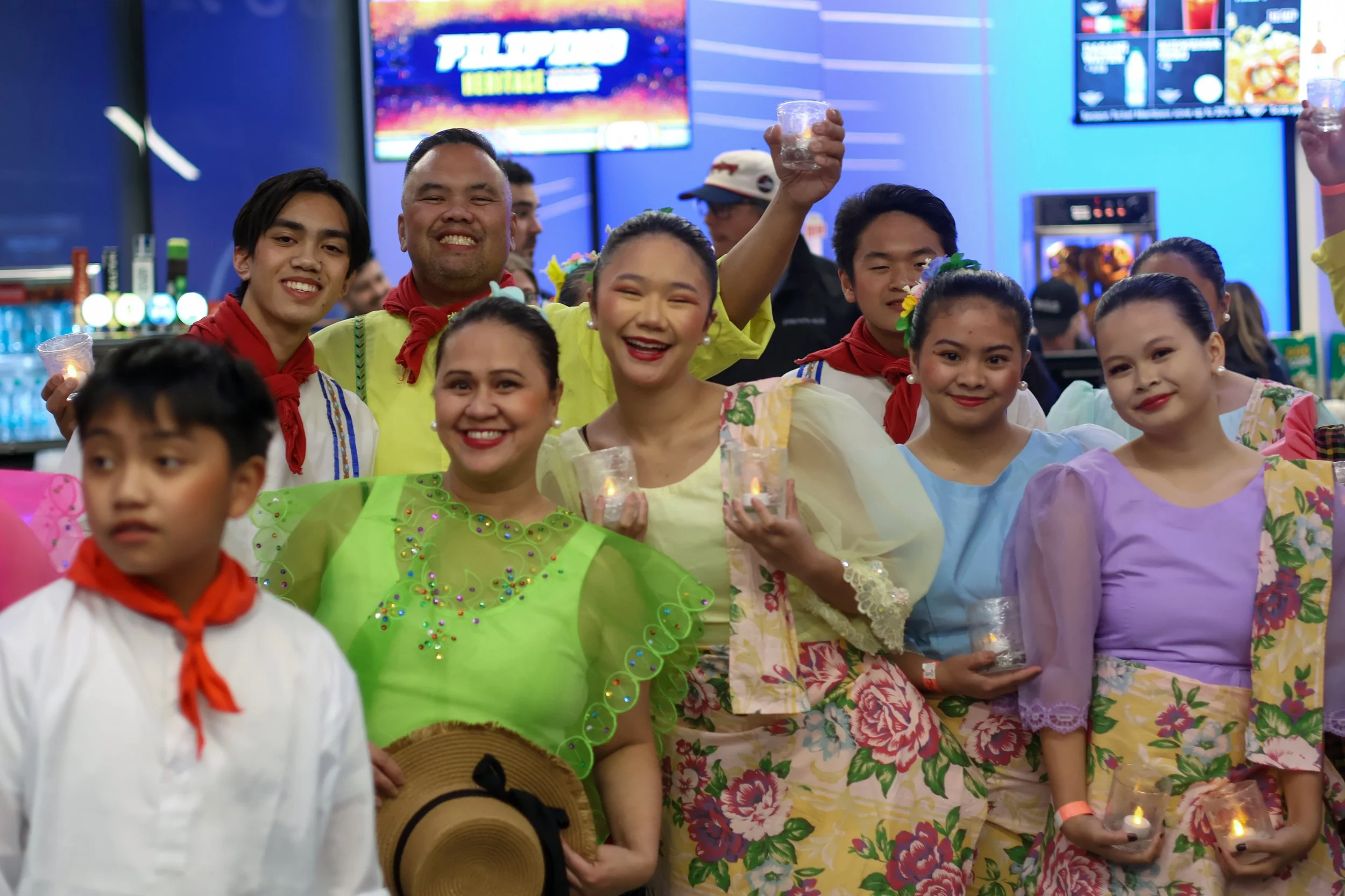 A group of people at a festive celebration, dressed in colorful traditional clothing, holding candles in small glasses, smiling and celebrating indoors.
