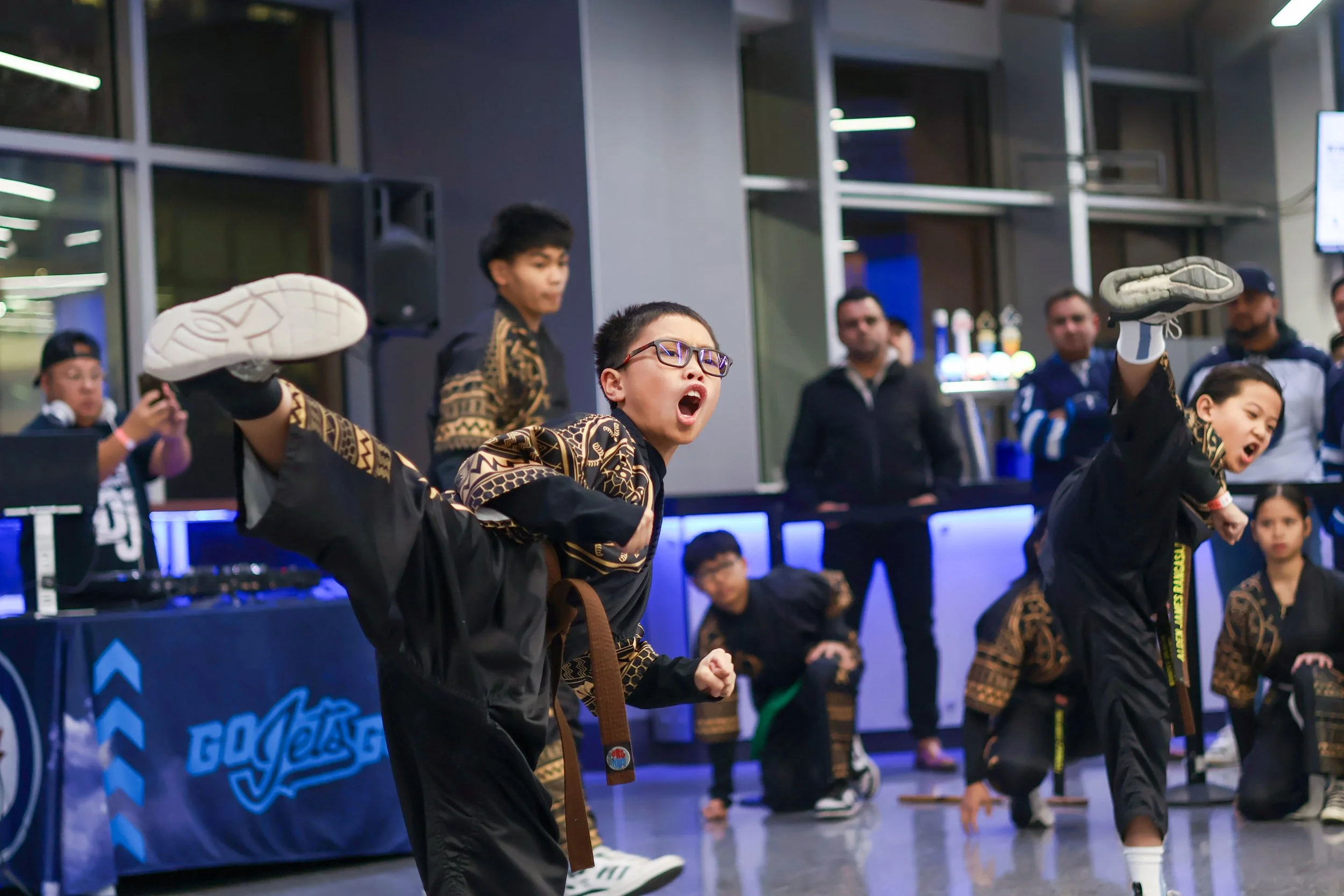 Children in martial arts uniforms performing high kicks in a martial arts demonstration while an audience watches indoors.