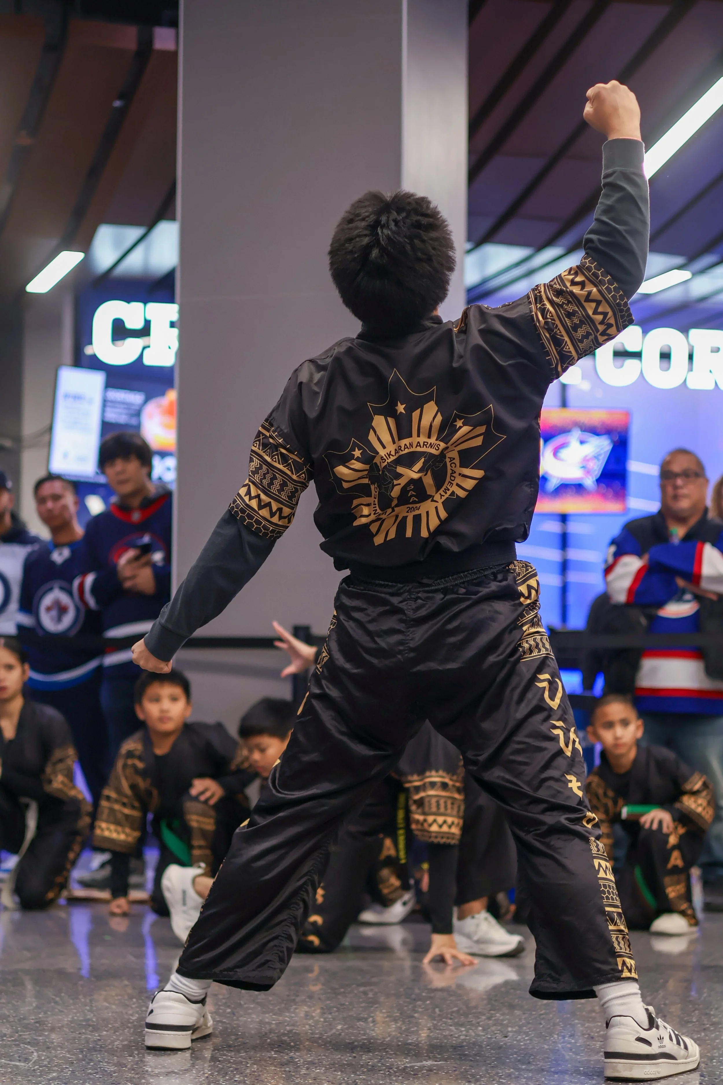A group of children dressed in martial arts uniforms, with one in the foreground striking a confident pose with his arm raised and back facing the camera. The background shows spectators and a digital screen.