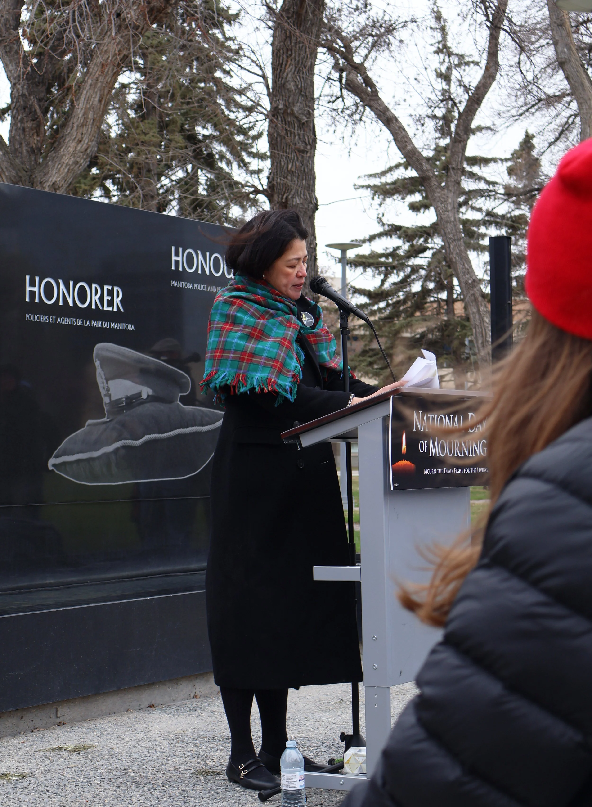 A woman at a podium reading aloud during a memorial event for the National Day of Mourning, with a large black sign behind her that displays an image of a moccasin and the text honoring Indigenous people.