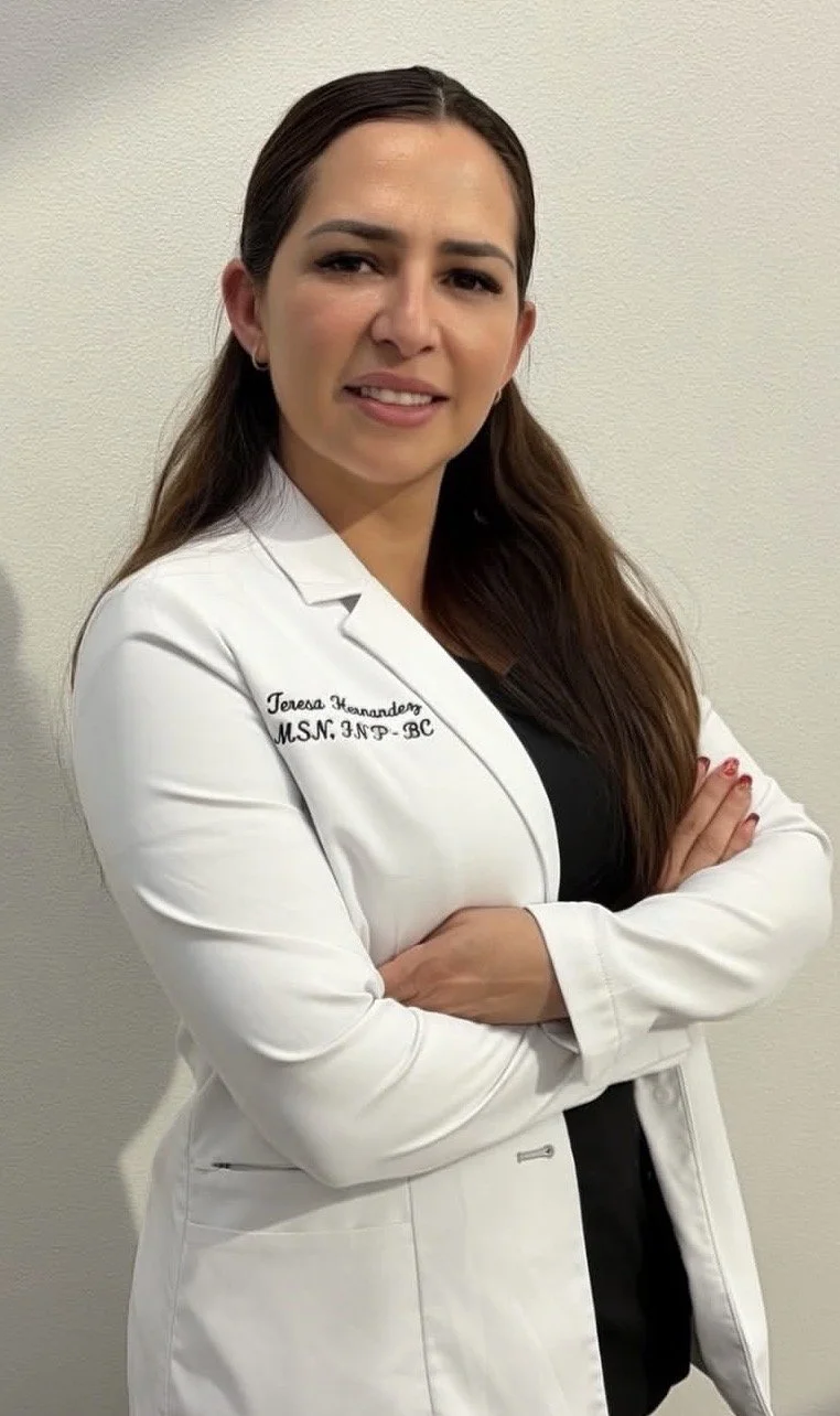 A woman in a white medical coat with her arms crossed, standing against a plain wall.