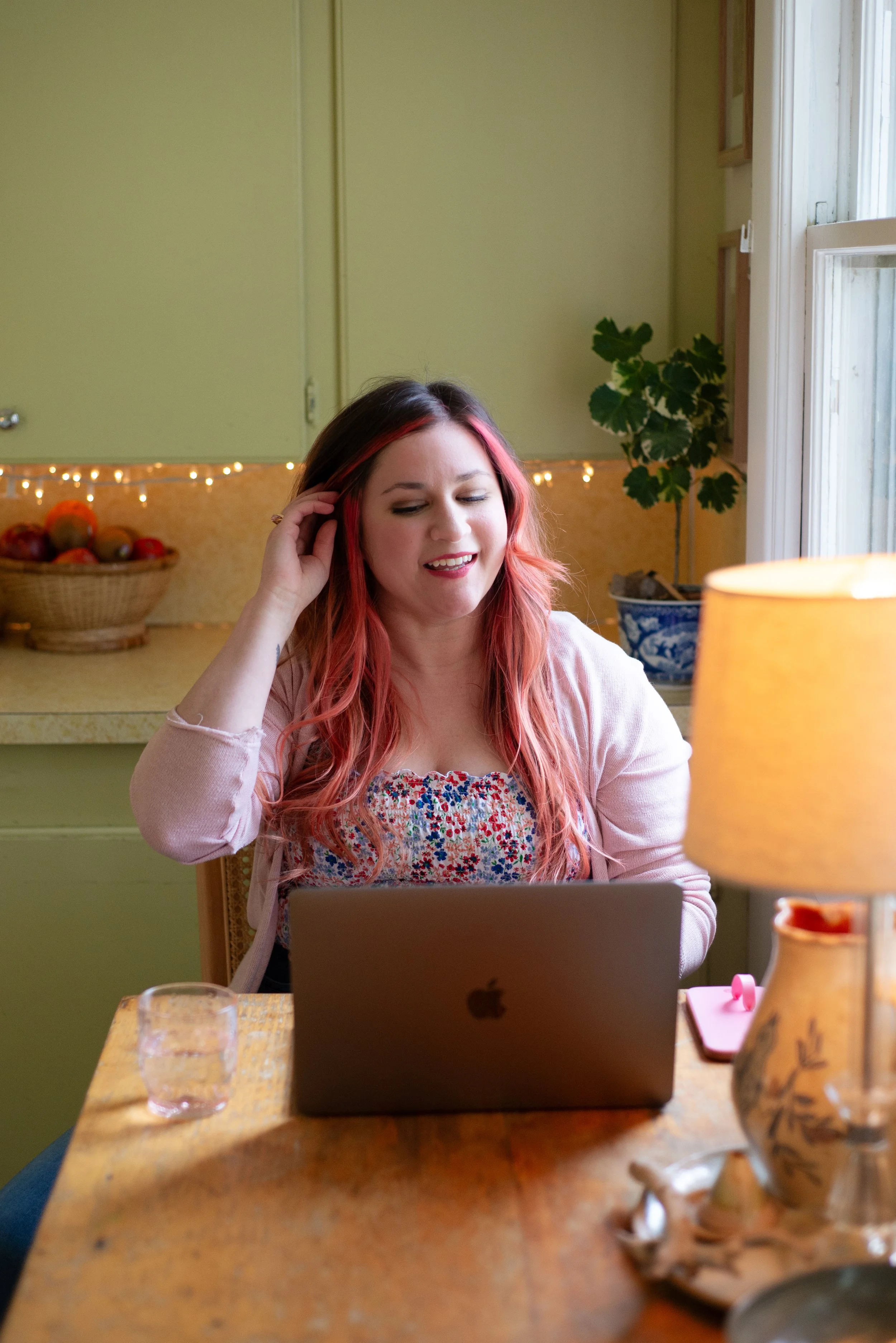 A woman with long, wavy red hair sitting at a wooden table, smiling at a laptop. She is wearing a pastel pink cardigan over a floral top, and appears to be in a cozy kitchen or dining area with green cabinets, a basket of fruit, and a potted plant in the background.