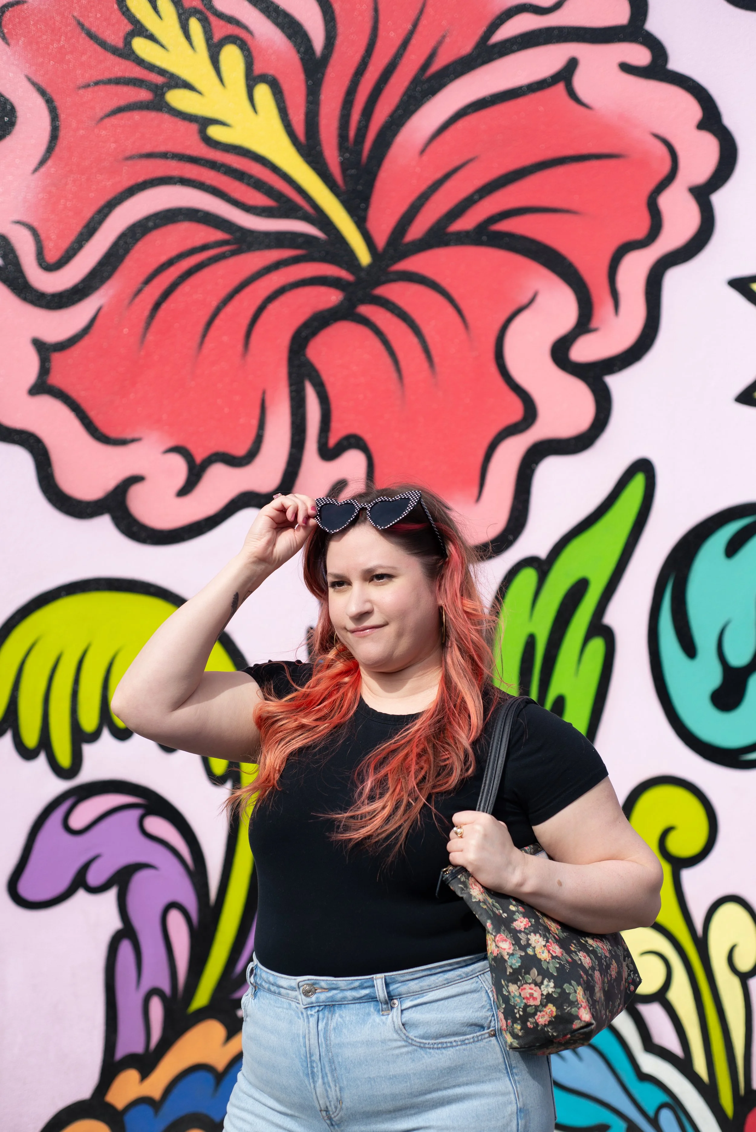 A woman with long, wavy red hair wearing a black t-shirt and light denim jeans, holding a floral-patterned bag over her shoulder and adjusting heart-shaped sunglasses while standing in front of a colorful mural featuring a large pink flower.